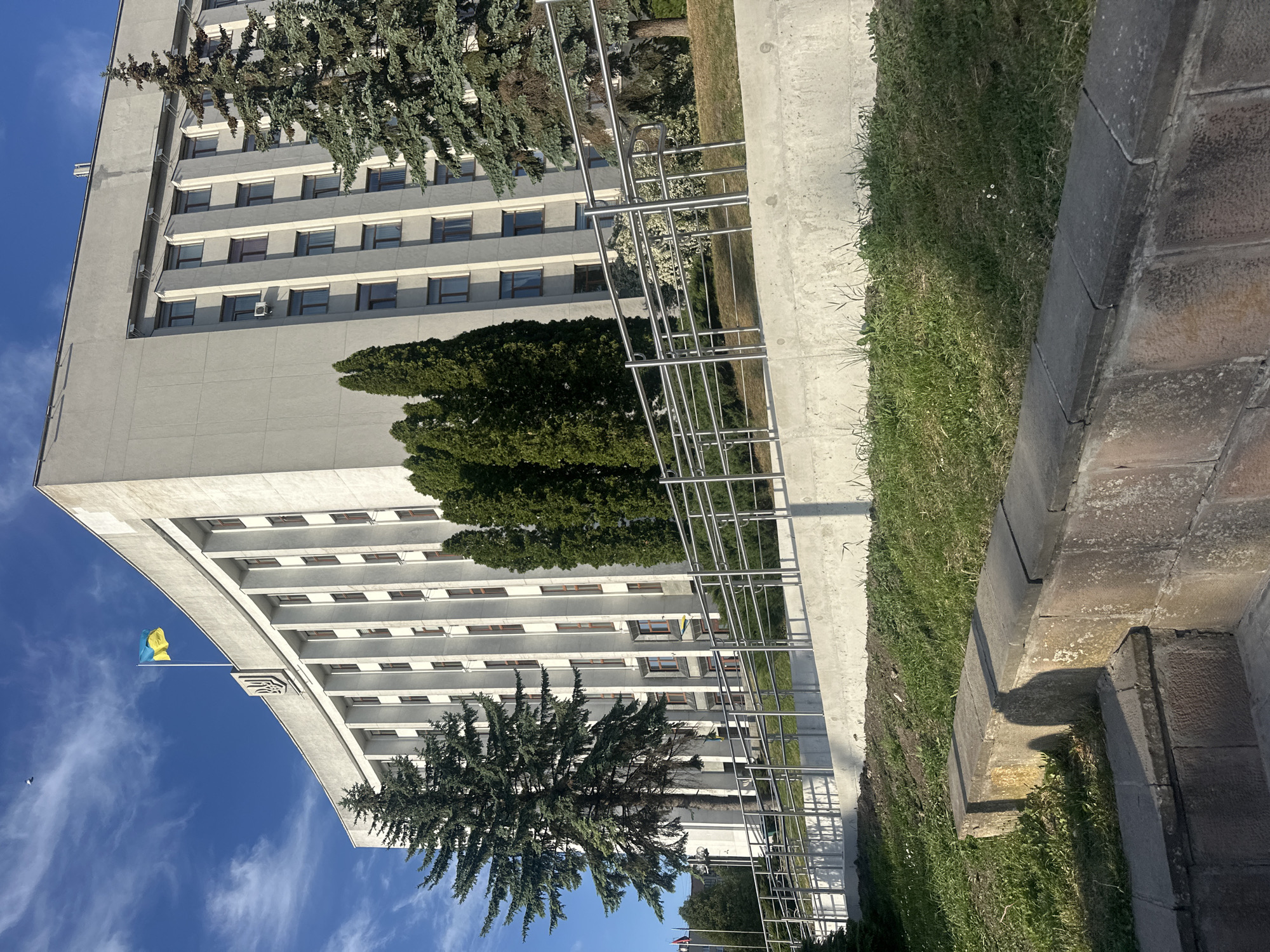 The image shows a view of the Ternopil Regional State Administration building in Ternopil, Ukraine. The gray, multi-story building features rows of windows and a curved facade with the Ukrainian flag visible against a blue sky with scattered clouds. A row of pine trees obscures some parts of the building. In the foreground, there is a stainless-steel structure and greenery including grass and stones.