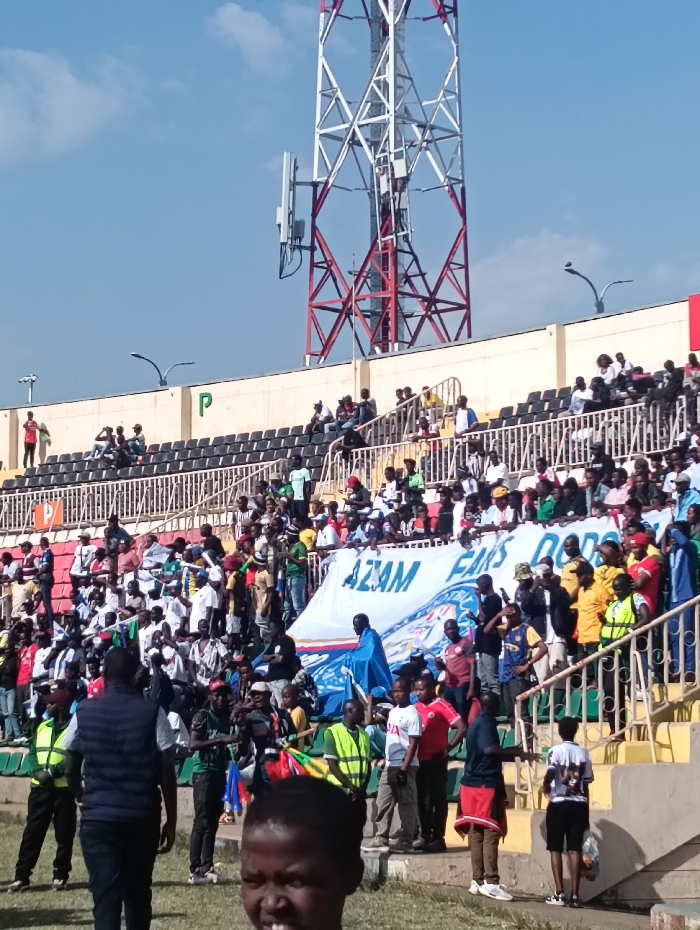 The image displays an outdoor stadium scene in Nairobi, Kenya, under a clear blue sky with minimal cloud cover. A prominent red and white lattice communication tower, equipped with multiple cellular antennas, extends high above the stadium structure in the background. The stadium features concrete tiers with rows of dark-colored seats, some occupied, some vacant. A beige wall with a green 'P' marking is visible on the upper left tier.
In the mid-ground, a large crowd, predominantly composed of male individuals appearing to be of African ethnicity, occupies the lower stadium stands and the grassy area adjacent to the field. Many are dressed in casual clothing, including t-shirts of various colors, while some wear bright yellow or green safety vests. A large white banner, held by multiple individuals, is visible, bearing blue text that reads "AZAM FANS DO..." and a circular logo. A person draped in a blue fabric stands near this banner. Various individuals are standing, observing, and some are holding small flags or other items.
In the immediate foreground, the head and shoulders of a young individual with dark skin and short dark hair are visible, partially obscuring the lower part of the scene. The individual faces slightly left and displays a slight smile. The ground directly in front of this individual is covered in grass.