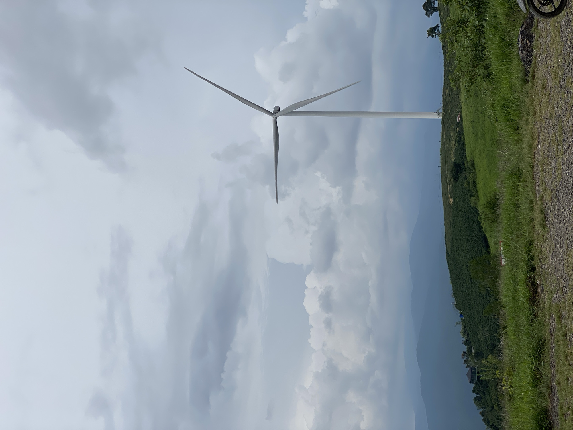 The image depicts a wind turbine standing tall on a grassy hillside in Trường Thọ, Vietnam. The sky is overcast with dramatic, gray clouds. In the foreground, a gravel path leads towards a lush green landscape dotted with trees and rolling hills. Several layers of distant mountains recede into the hazy horizon. A portion of a motorcycle is visible in the lower right corner of the frame.