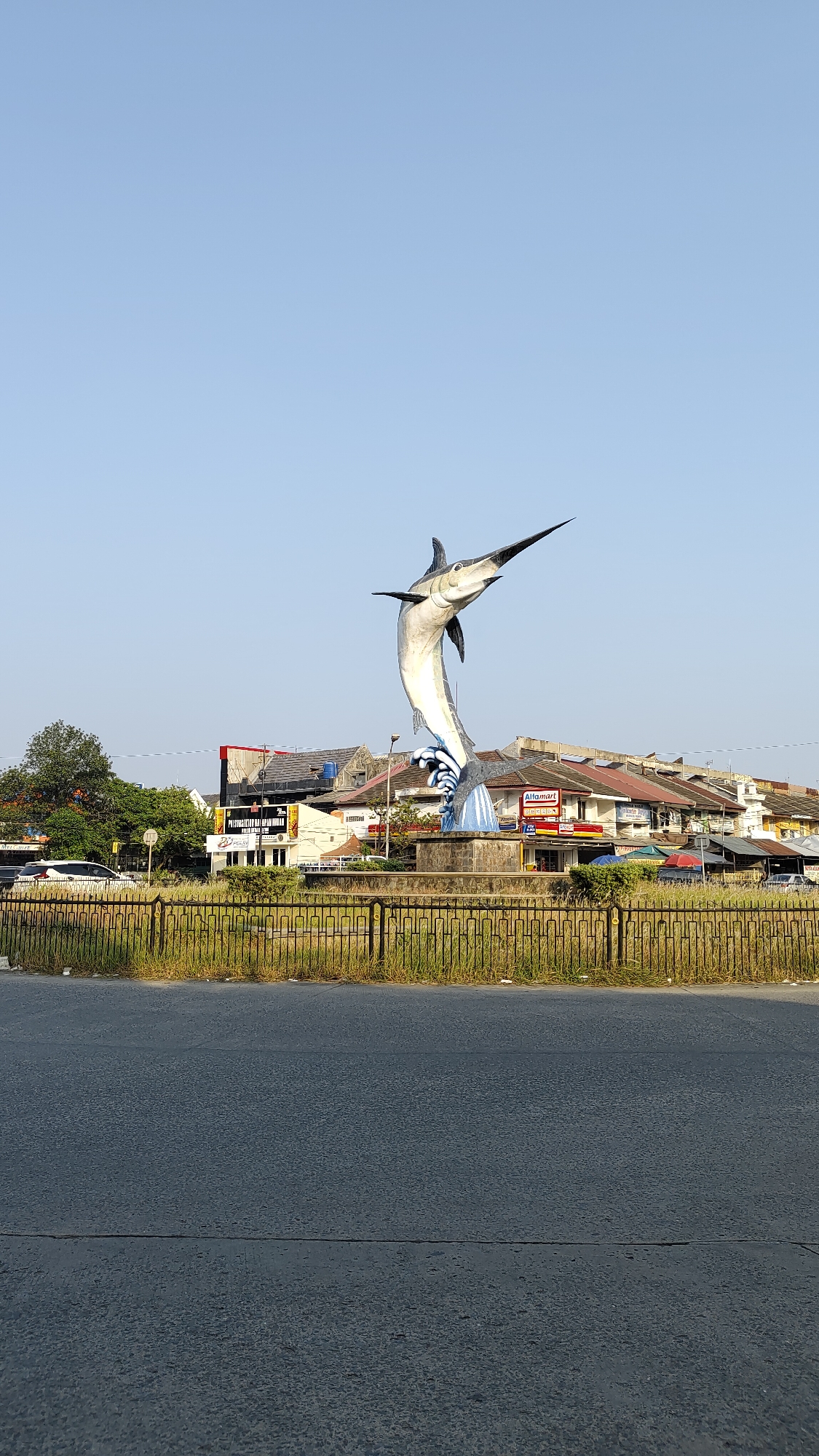 A tall, blue and silver marlin statue stands on a concrete base in a roundabout in Bekasi, Indonesia. The statue is angled upwards, with its mouth open and its bill pointed to the sky. It appears to be leaping out of stylized blue waves. Behind the statue, there are several buildings, including a red-roofed building with a sign for "Alfamart" and another building with a sign that says "Peringati Ulang Tahun." A black metal fence runs along the edge of the roundabout, with dry grass and some litter visible behind it. The statue is positioned in the middle of a road, with asphalt visible in the foreground and a clear blue sky overhead. A few trees are visible to the left of the statue, and some cars are parked or driving on the road.