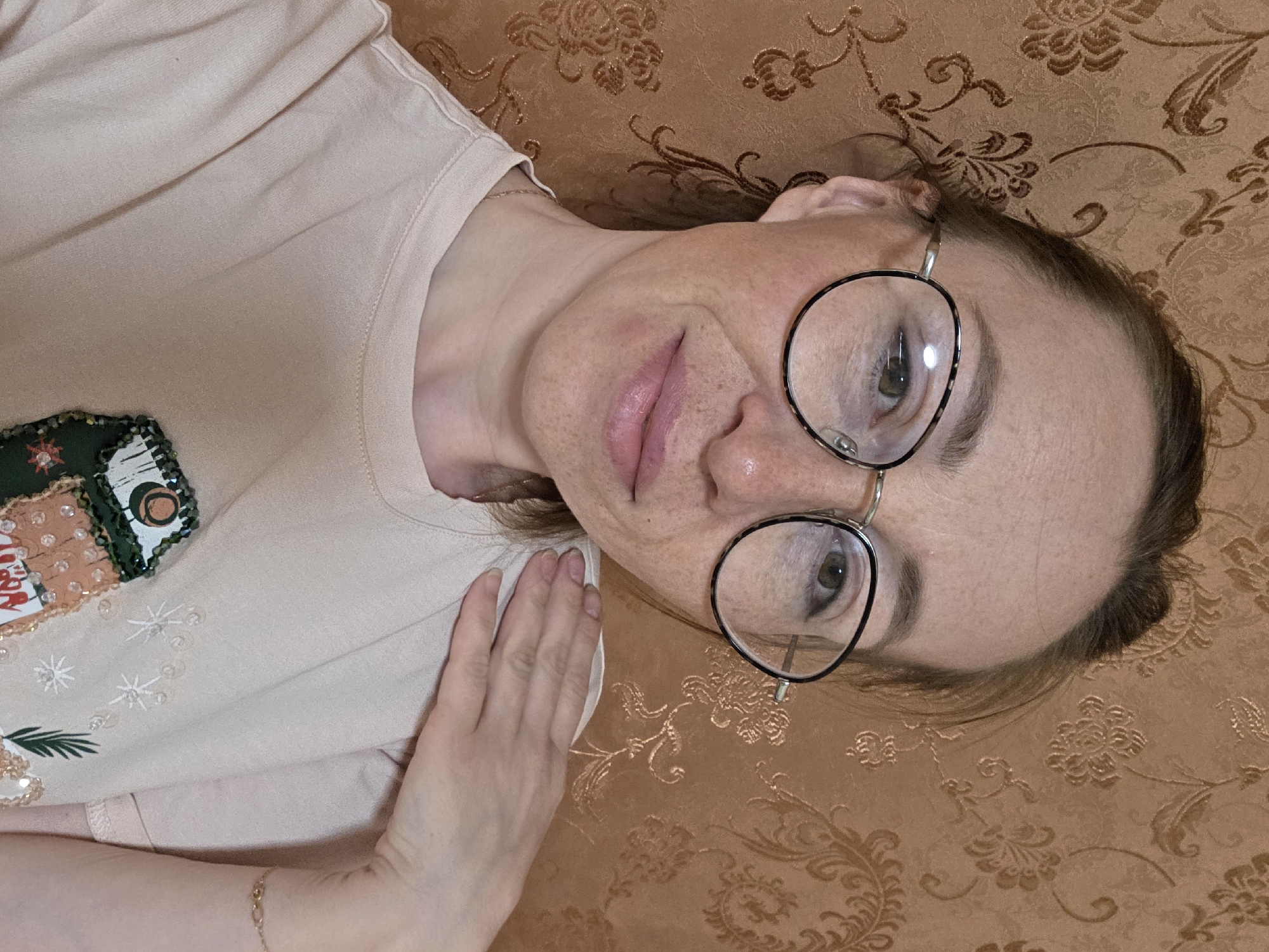 The image displays a close-up, head-and-shoulders portrait of an adult female, looking directly into the camera. She has fair skin with visible freckles, brown hair pulled back with some strands framing her face, and wears round eyeglasses with thin gold frames and dark, possibly tortoiseshell, rims. Her eyes are greenish-brown, and her lips are light pink with a slight upward curve. A thin gold chain necklace is visible around her neck. She is wearing a light peach-colored crew-neck t-shirt featuring a partially visible, embellished design on the left side of the chest, which includes elements resembling a house, trees, and snowflake-like patterns, adorned with beads and sequins. Her right hand, displaying natural nails, is positioned on her left shoulder. A thin gold chain bracelet is visible on her left wrist. The background is an indoor wall covered in brown or tan wallpaper with a darker, ornate floral and swirling pattern. The specific location within the city of Chaikovsky, Russia, is not discernible from the visual information provided in the image.