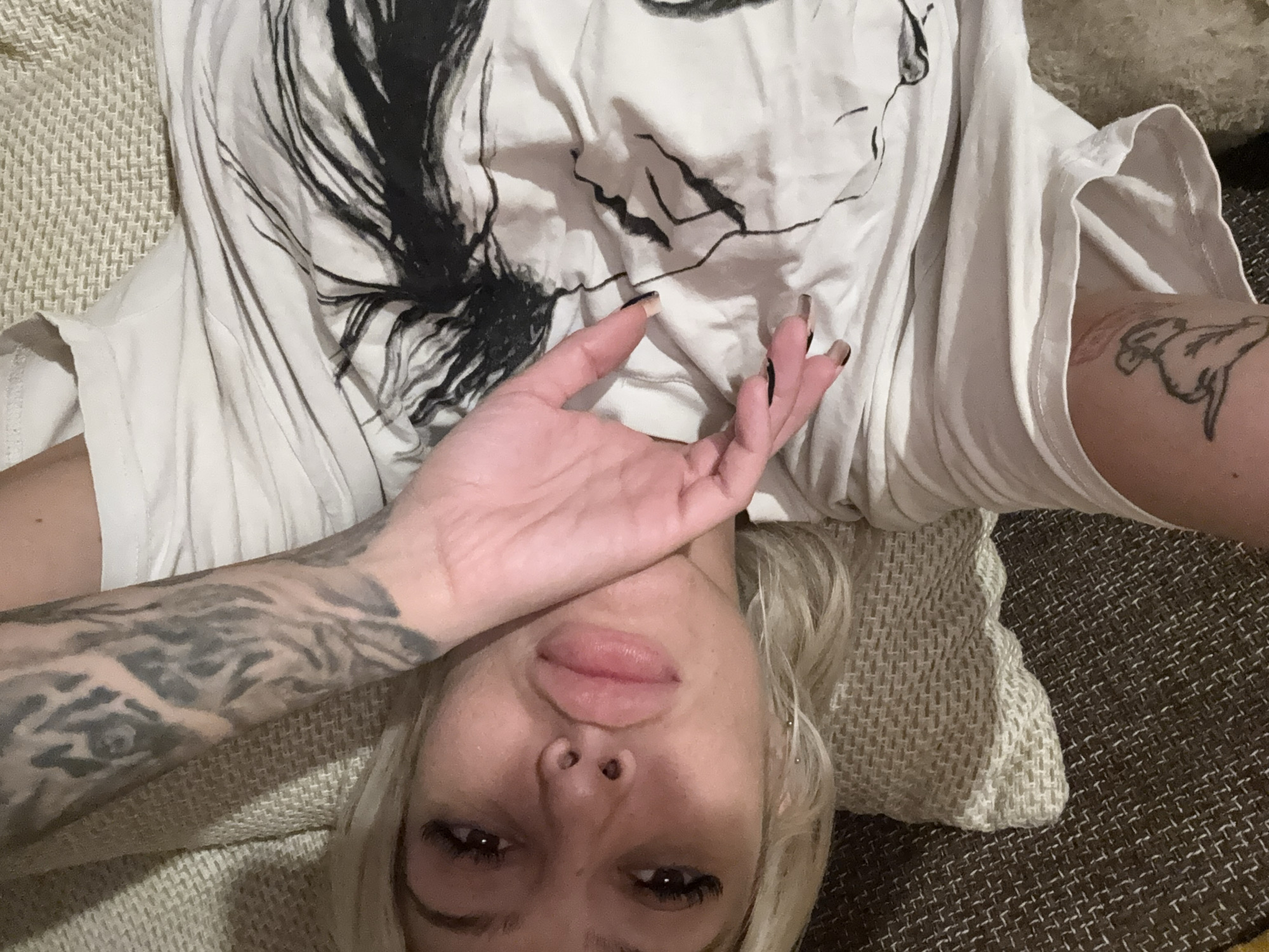 This media file features a young woman lying down in an indoor setting, likely on a couch or bed in Plovdiv, Bulgaria. The image is taken from an overhead perspective, possibly a selfie.
**Subjects:** The central subject is a woman with blonde hair and dark eyes. Her right hand, adorned with dark nail polish, is raised and partially covers her forehead and eyes. She has tattoos on both arms: a full sleeve of intricate dark designs is visible on her left arm, and a linear, stylized tattoo (possibly depicting an animal or symbol) is on her right bicep. She has full lips, a subtle nose piercing, and visible eyelashes.
**Objects:** She is wearing a white t-shirt featuring a large, abstract dark graphic design that appears to be a stylized face or feathered pattern across the chest area. She is resting on a light-colored, textured knitted fabric, which lies on a darker, tightly woven surface, suggesting bedding or upholstery.
**Activity or event taking place:** The woman is captured in a relaxed, prone position, looking directly up towards the camera. Her pose, with a hand partially obscuring her face, gives the impression of a candid or intimate moment, perhaps one of quiet contemplation or slight weariness.
**Notable details:** Her expression suggests a relaxed, possibly pensive or slightly pouting mood, with her lips slightly parted. The indoor lighting is soft, not indicating a specific time of day but suggesting a comfortable, personal space. There is no visible text in the image.