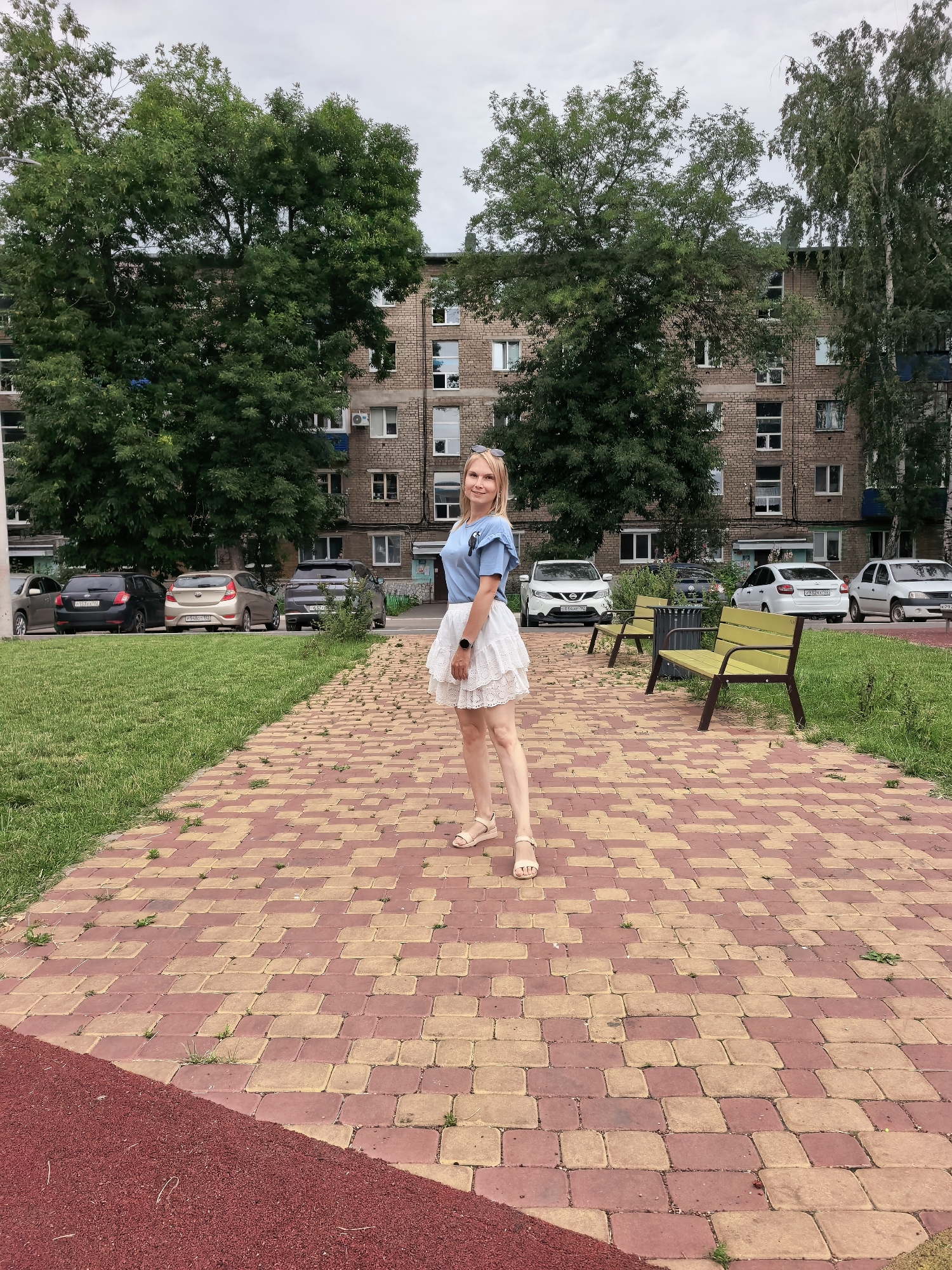 A full-body, medium shot captures a blonde woman standing on a paved walkway in an outdoor setting. She is wearing a blue t-shirt with a graphic print on the left chest and a white, ruffled skirt. Her feet are adorned with tan sandals. The walkway is made of rectangular red and tan paving stones, and a section of red ground cover is visible in the lower left corner. Behind her, a line of parked cars is visible on a street adjacent to a green lawn. In the background, a multi-story brick apartment building stands under a cloudy sky, with several mature trees flanking it. The scene appears to be a residential area in Sterlitamak, Russia.