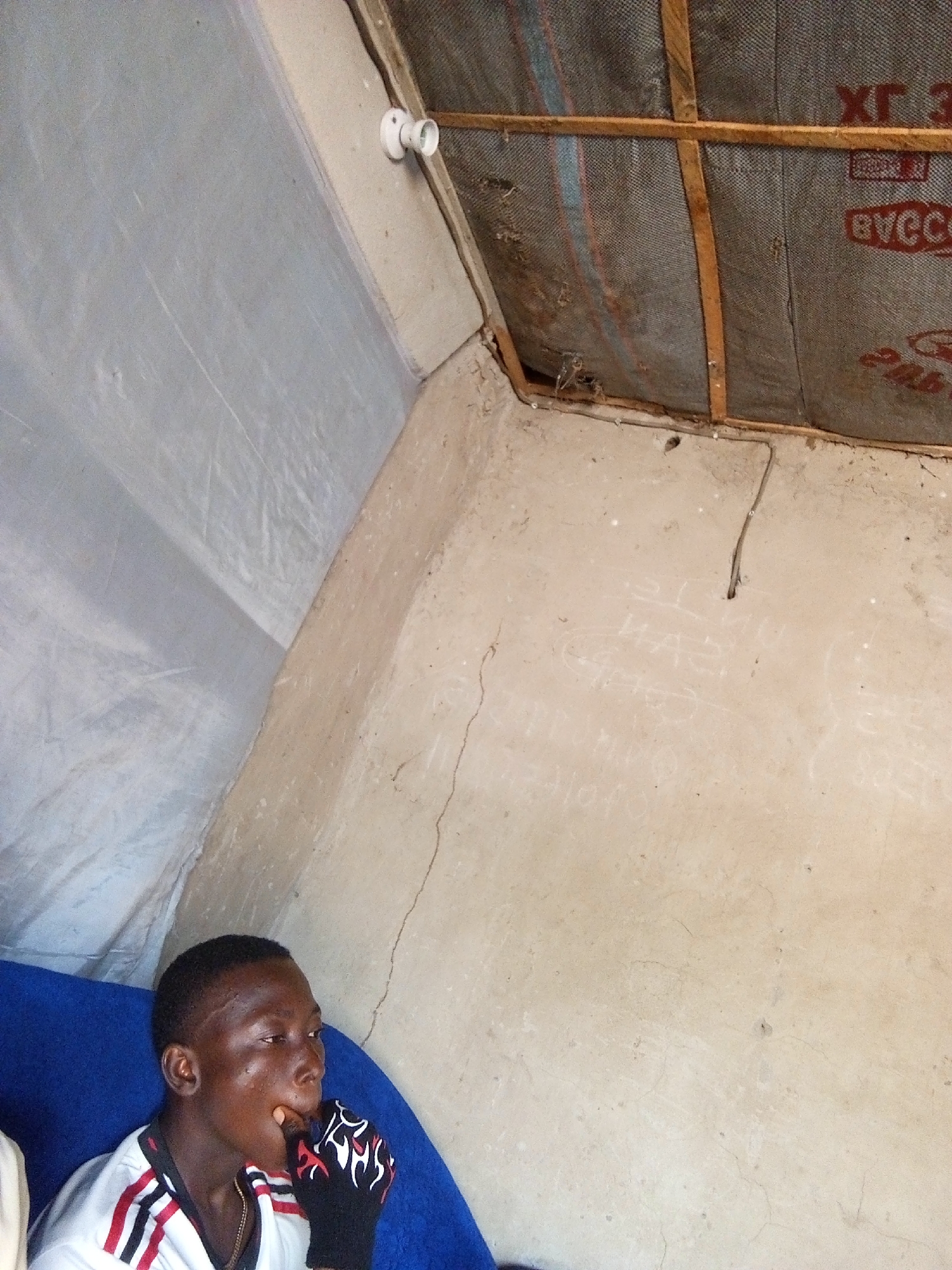 This image depicts a boy sitting on a blue surface in what appears to be a rustic indoor environment. The walls are a light, earthy color, with some visible cracks and scribbled markings. Above, the ceiling is covered with a textured material, possibly insulation, supported by wooden beams. A bare light socket is visible on the ceiling. The boy is wearing a white t-shirt with red and black stripes on the collar and sleeves, and a black glove with a flame-like pattern on his left hand, which he is using to touch his mouth. He is looking to his right, with his mouth slightly open. The overall lighting suggests it is daytime, with the light source appearing to be from the left. The scene has a simple and possibly makeshift structure, suggesting it could be a temporary shelter or dwelling in a less developed area. Without further information, it is impossible to determine the exact location within Nyabe, Nigeria, other than it is an indoor setting.