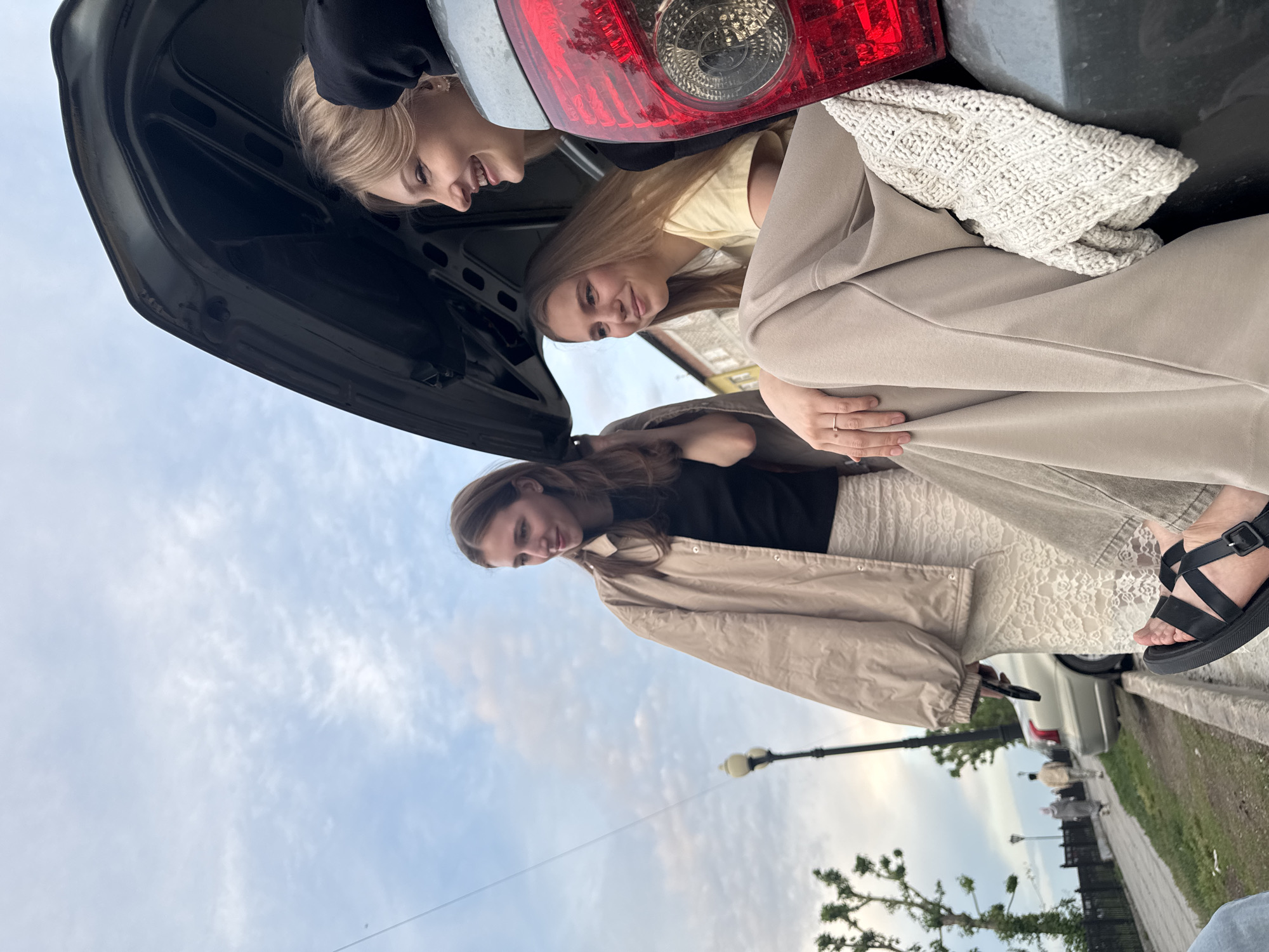 Three young women are looking into the open trunk of a gray car. The trunk lid is open, with a red taillight visible. One woman has blonde hair and is wearing a black top. Another woman is wearing a yellow top. The third woman has brown hair and is wearing a black top with a tan jacket and a white lace skirt. She is also wearing black sandals and is holding a phone. The background shows a city street with a lamppost, trees, and a sidewalk. Other pedestrians are visible in the distance. The sky is partly cloudy. The location is Perm, Russia.