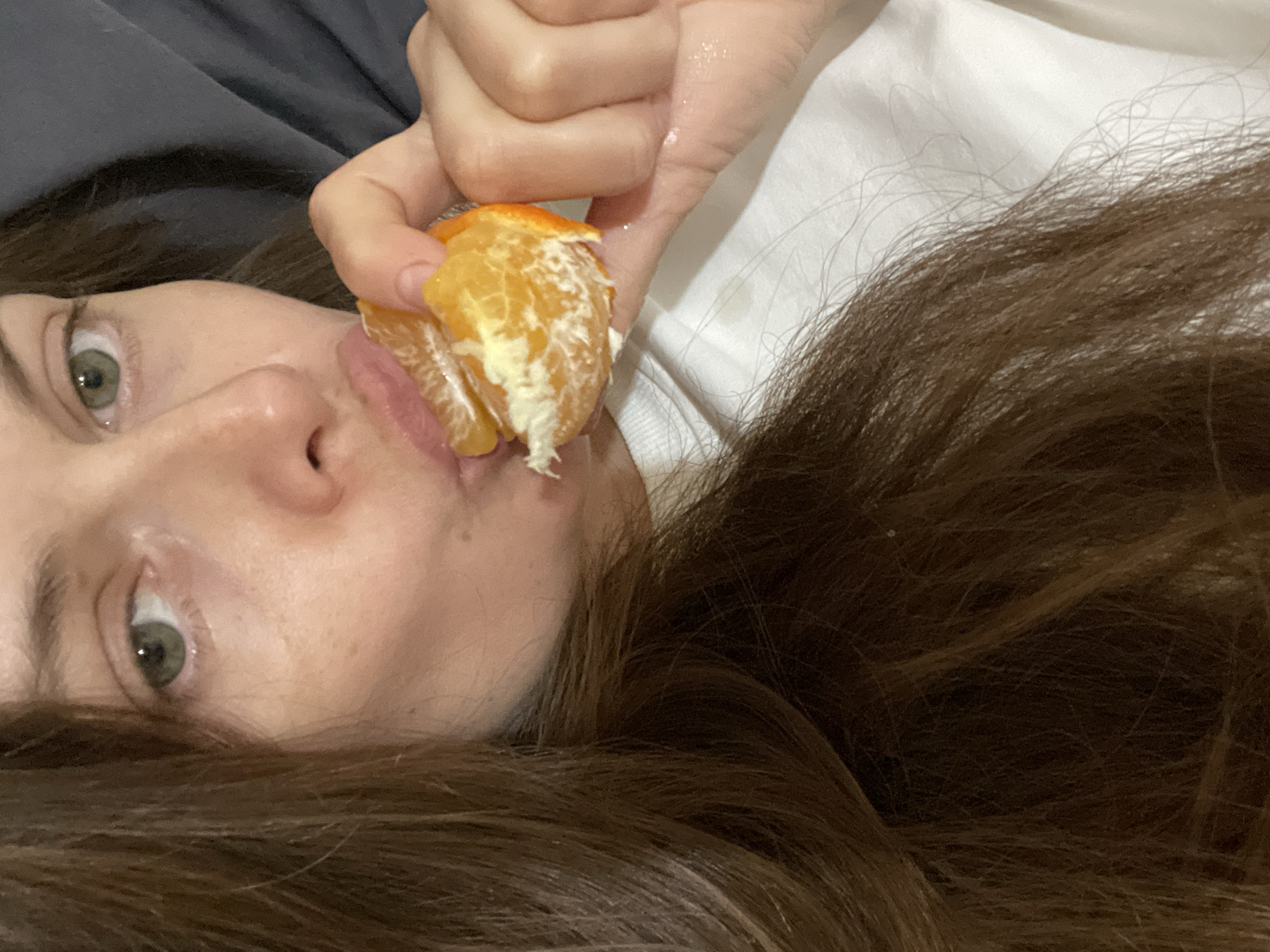 A close-up shot shows a person's face, from the eyes down to their chest, as they eat a peeled tangerine. The person has light skin and long, brown hair that frames their face. They are wearing a white t-shirt. The background is dark and out of focus, possibly a blanket or pillow. The image does not provide enough information to determine a specific location within the city of Заречный, Russia.