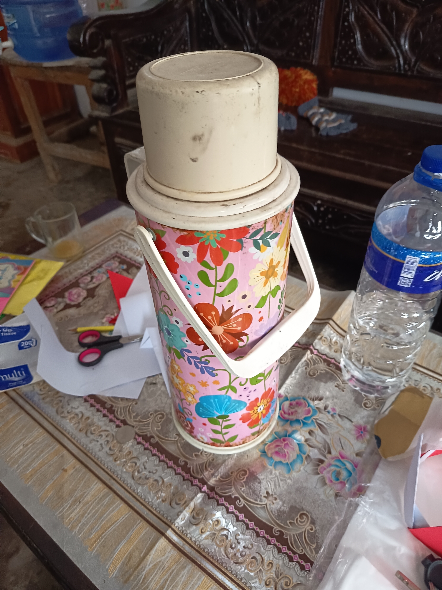 A close-up shot focuses on a vintage floral thermos on a table with a patterned tablecloth. To the right of the thermos, a partially filled plastic bottle of water with a blue label is visible. In the background, parts of a wooden chair are seen. Scattered on the table are various items including scissors, a pencil, papers, and a glass mug with liquid. The scene appears to be an indoor setting, possibly a home in Krampilan, Indonesia, given the presence of common household items and the style of the furniture.