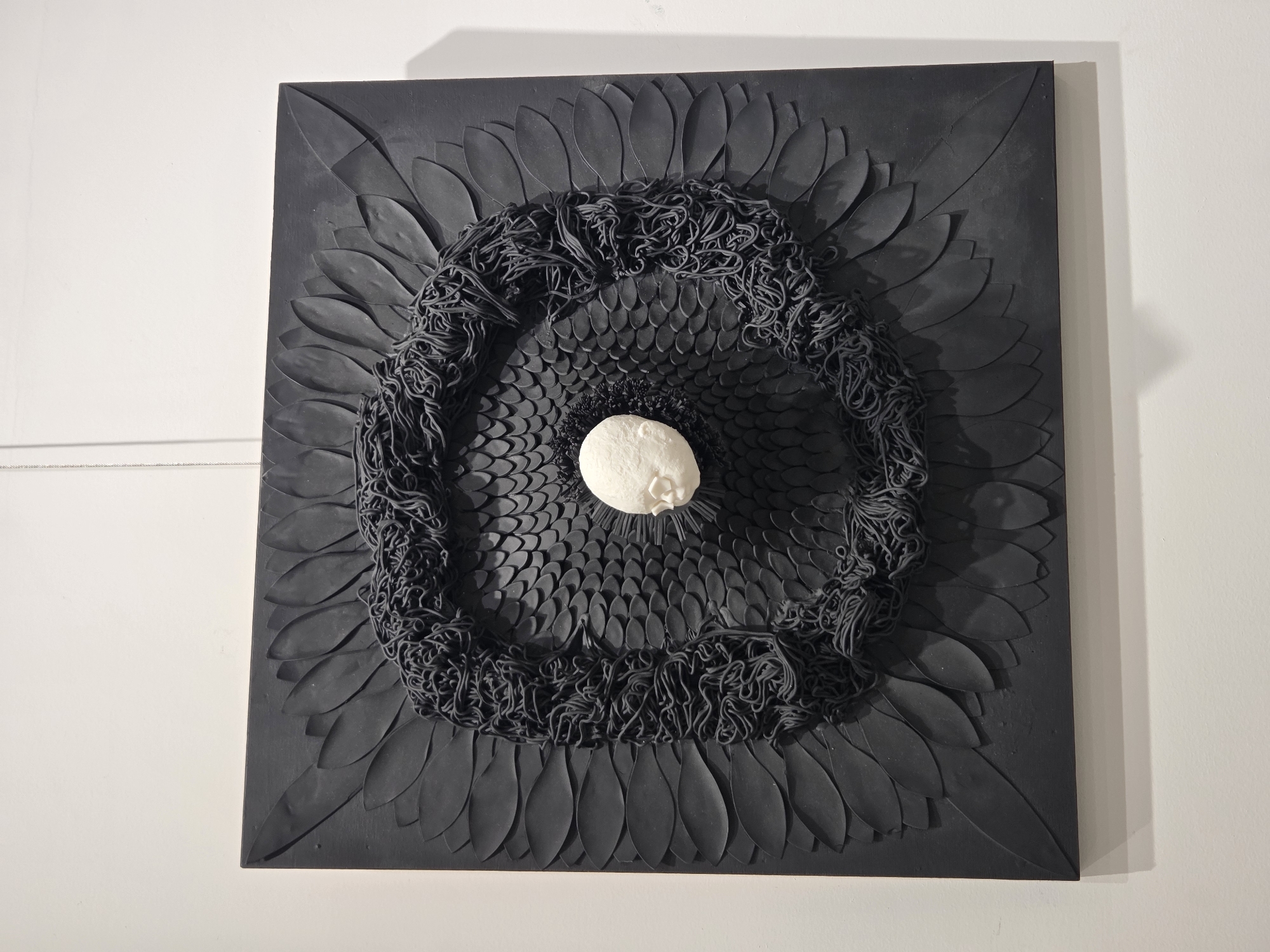 This is a close-up, eye-level, full-shot photograph of a black, square, framed artwork hanging on a white wall. The artwork is a mixed-media piece with a strong central focus and intricate details. The background of the artwork is a flat black square. Layered on this square are two distinct textures. The outer perimeter of the artwork features a series of dark, petal-like shapes radiating outwards, creating a floral or feathered effect. Inside this outer layer, the texture shifts to a pattern of small, overlapping, scale-like shapes, arranged in concentric circles that form a concave recess towards the center.
Dominating the center of the artwork is a circular arrangement of thick, black, string-like material, coiled and tangled into a dense, almost nest-like form. This coiled material creates a dark, textured vortex. Embedded within the very center of this coiled mass is a small, white, three-dimensional sculpture of a human head, facing slightly to the right. The head appears to be made of a smooth, pale material, possibly ceramic or plaster. The contrast between the stark white of the head and the overwhelming black of the artwork is striking.
The lighting in the photograph casts a subtle shadow from the artwork onto the white wall below it. A thin, white wire is visible extending from the top of the artwork, suggesting it is suspended or mounted. The image appears to have been taken in an indoor gallery or exhibition space, given the clean white walls and professional presentation of the artwork. There are no people or other distracting elements visible in the frame.