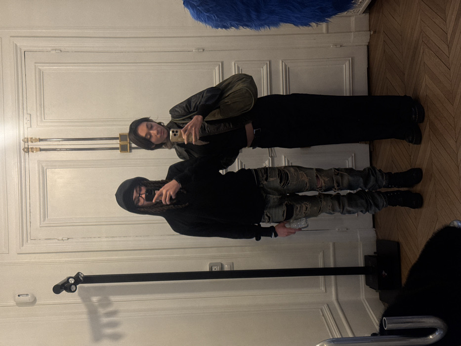 The scene depicts two individuals taking a mirror selfie in an interior space characteristic of a Parisian apartment, featuring white walls with classical molding, tall ornate white double doors with gilded hardware, and a light-toned herringbone parquet floor.
On the left, a male individual wears a black hooded sweatshirt, dark-rimmed glasses, and heavily distressed light-wash jeans with patches. He has long dreadlocks or braids, holds a clear plastic water bottle in his left hand, and a gold-cased smartphone in his right, angled towards the mirror. He wears black boots.
On the right, a female individual wears a dark cropped top, a black leather jacket, and a long black skirt, complemented by black platform shoes. She carries an olive-green hobo-style shoulder bag and holds a gold-cased smartphone in her right hand, also directed towards the mirror. A light-colored scarf or accessory is visible around her neck.
To the left of the male, a tall black freestanding spotlight fixture with three visible lights stands near a white wall outlet. In the lower-left foreground, a segment of a dark, textured object is visible. On the far right, a large, vibrant blue, shaggy object is partially captured. Both individuals are focused on their respective phones or reflections, engaging in the act of photography.