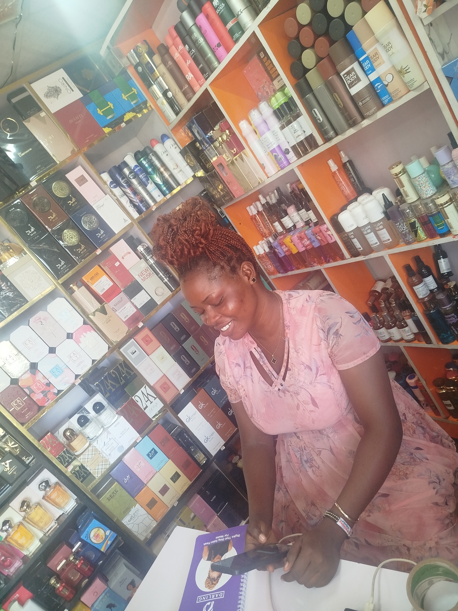 The image depicts a woman with braided, reddish-brown hair and a light pink floral dress smiling as she looks down at a counter filled with a diverse collection of perfumes and body sprays. The shelves behind her are also packed with various beauty products, including deodorants, lotions, and more fragrances. The sheer volume and variety of items suggest a retail setting, likely a perfume or cosmetics store.
Visible text includes brand names and product descriptions such as "DARLING," "DEAR BODY," "NIVEA," "GK men," "ASAD," "AVANTI," "ONIRO," and "CONFESSION." Some of the text is in Arabic script, indicating the international nature of the products. The lighting in the store appears bright, suggesting it is daytime. The woman's attire and the abundance of products create a lively and perhaps busy atmosphere. The woman is holding a black electronic device in her right hand and appears to be engaged in some activity related to the products on the counter. The overall impression is one of a well-stocked store catering to personal care and beauty, possibly in a bustling commercial area in Ugbowo, Nigeria.