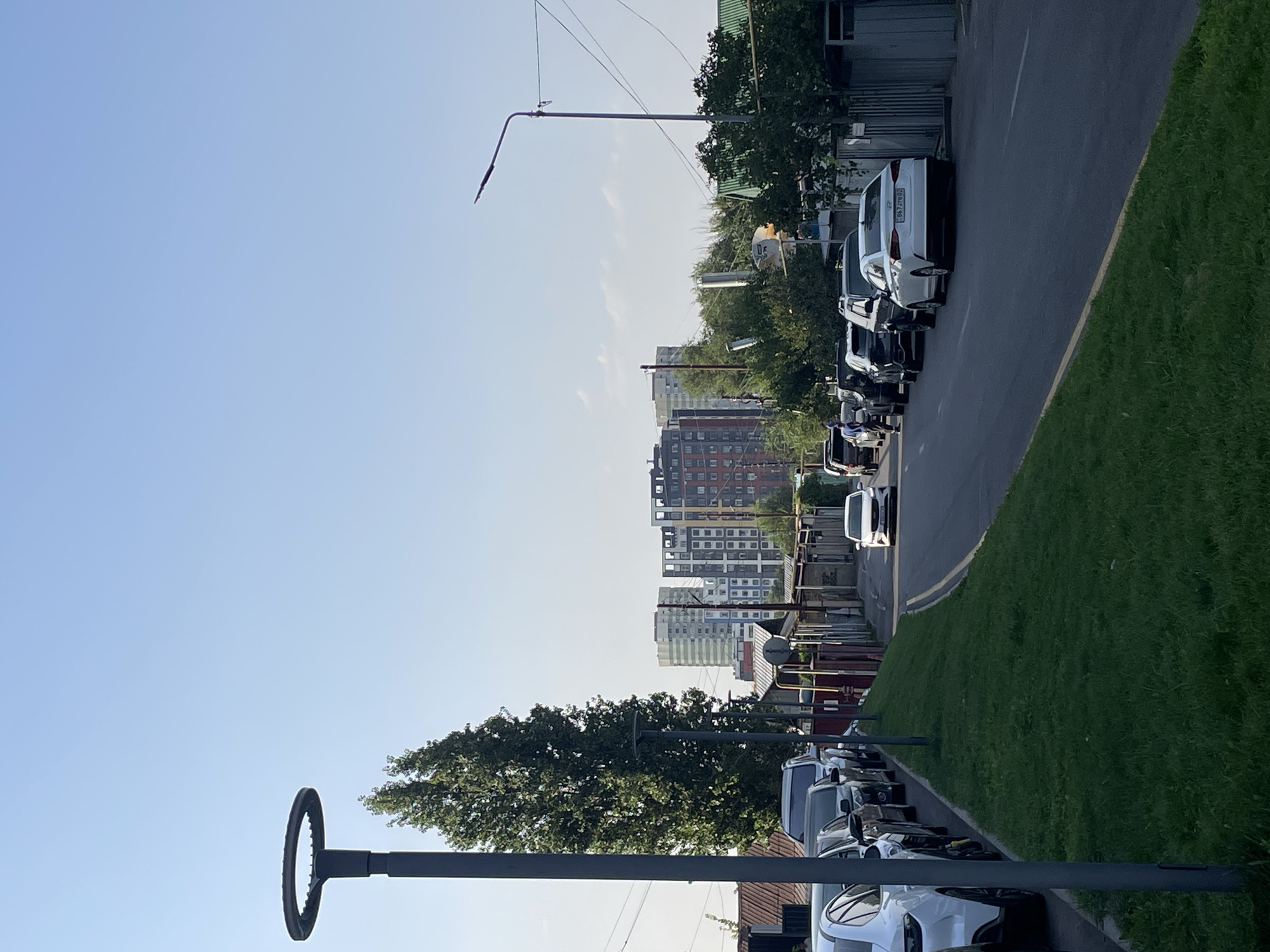The image depicts a street scene in Almaty, Kazakhstan, characterized by residential buildings and parked vehicles. A tall, modern street lamp with a circular top stands prominently on the left side of the frame. Adjacent to it is a large, mature tree with dense green foliage. The street is lined with parked cars on both sides, including a white sedan in the foreground on the left and a white sedan further down the street on the right. Several other vehicles, varying in color and type, are also visible, suggesting a residential area. In the background, multi-story apartment buildings with distinct architectural styles and colors rise against a clear blue sky. The environment appears to be a typical urban street with a mix of older and newer constructions, with some greenery interspersed amongst the buildings. The overall impression is one of a sunny day in a developed urban area.
