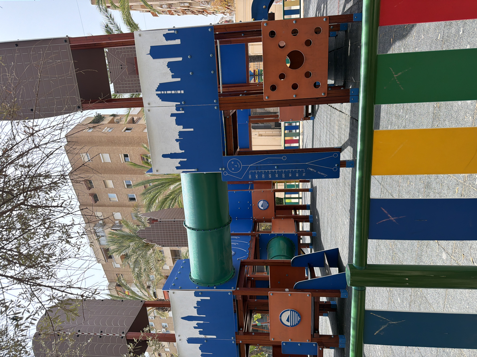This is a medium shot, taken outdoors, of a colorful children's playground structure. The playground appears to be constructed from a variety of materials, including wood, metal, and plastic. It features several different levels and sections, including a large green tube, what looks like a slide, and various panels with decorative elements. One panel has a blue silhouette of a city skyline. Another panel is orange with circular cutouts and a circular graphic inside. There is also a ruler-like marking on a blue panel that appears to be a height chart, with numbers from 70 to 150.
In the background, there are buildings, some of which appear to be apartment complexes. Palm trees and other foliage are also visible, suggesting a warm climate. The ground beneath the playground is paved with a gray, textured material. The sky is visible in the upper portion of the image and appears to be clear and bright, suggesting it is daytime with fair weather.
There are no people visible in the image, so no specific activity or event is being captured, other than the presence of the playground itself. Notable details include the vibrant colors of the playground elements—red, green, yellow, and blue—contrasting with the more muted tones of the surrounding buildings and foliage. The overall impression is that of a public space designed for children's recreation. There is some visible graffiti or markings on some of the blue panels of the structure. The visible text includes numbers and what appears to be a question in Spanish: "Cuanto mides?".