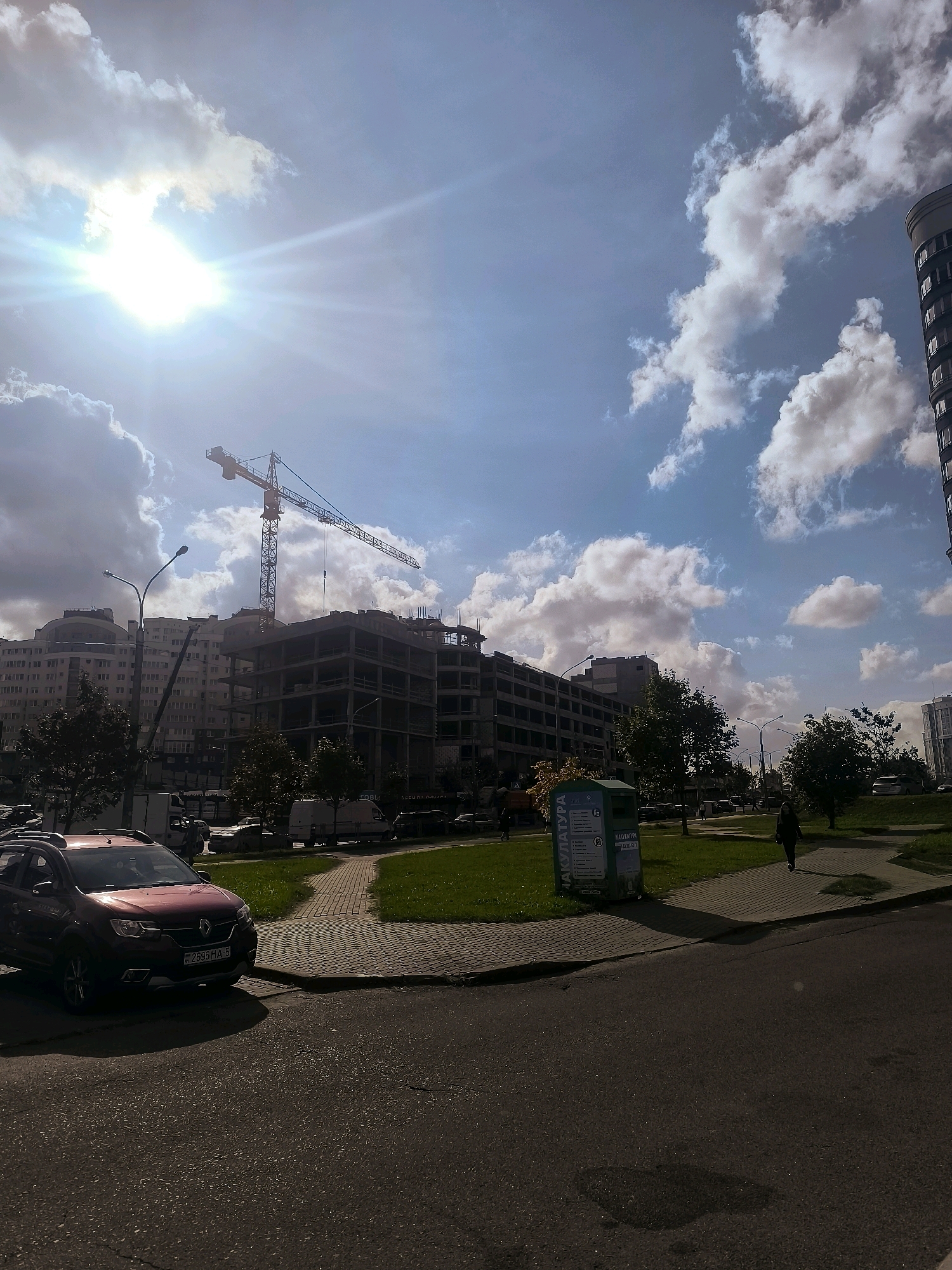 The image depicts a construction site in Minsk, Belarus, on a sunny day with scattered clouds. A large yellow crane dominates the skyline over a multi-story building under construction, showing exposed concrete framework. Adjacent to the construction site, a completed residential building with numerous windows is visible. In the foreground, a red SUV is parked on a paved surface, and a pathway leads towards a green area with trees and a blue recycling bin. A woman walks along the pathway in the mid-ground. The overall scene suggests an urban environment undergoing development.