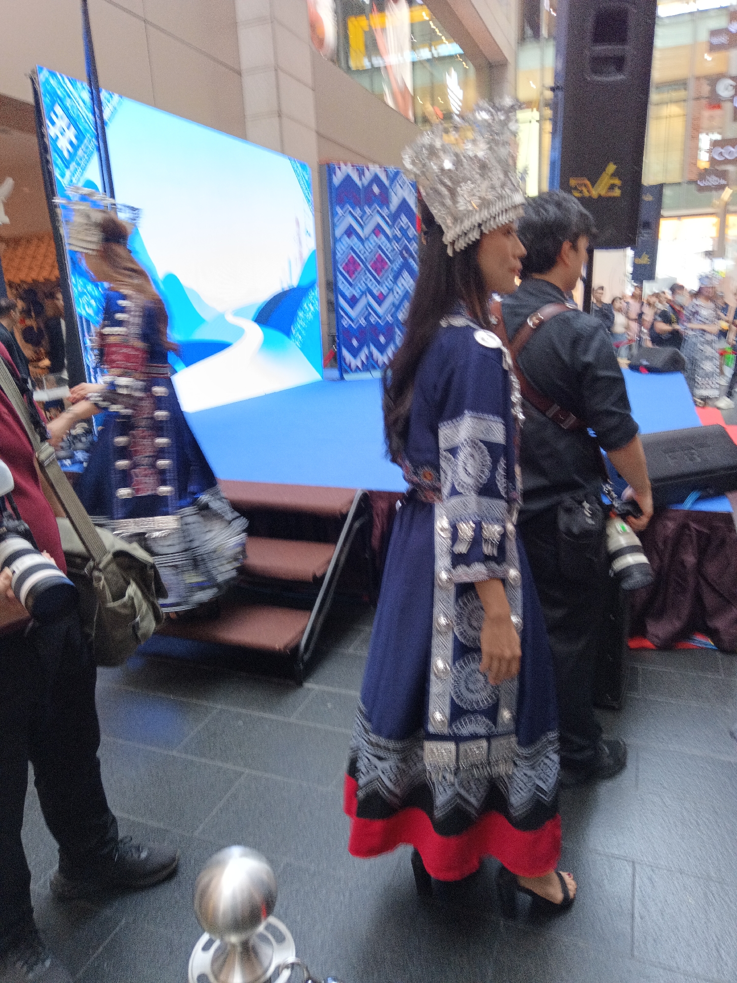 In a public indoor space, possibly a shopping mall, a woman in traditional attire, featuring a silver headdress and a dark blue dress with intricate silver embroidery and a red hem, is standing. She is facing towards the right of the frame, her body angled away from the camera. Behind her, a large screen displays a blue abstract landscape with a white path. To her right, a man in a black shirt and pants is visible, with a camera strap across his shoulder and a camera in his hand. Several other individuals are present in the background, some appearing to be spectators. The ground is paved with dark tiles. The overall lighting suggests an indoor, well-lit environment, typical of a commercial or exhibition area in Kuala Lumpur, Malaysia.