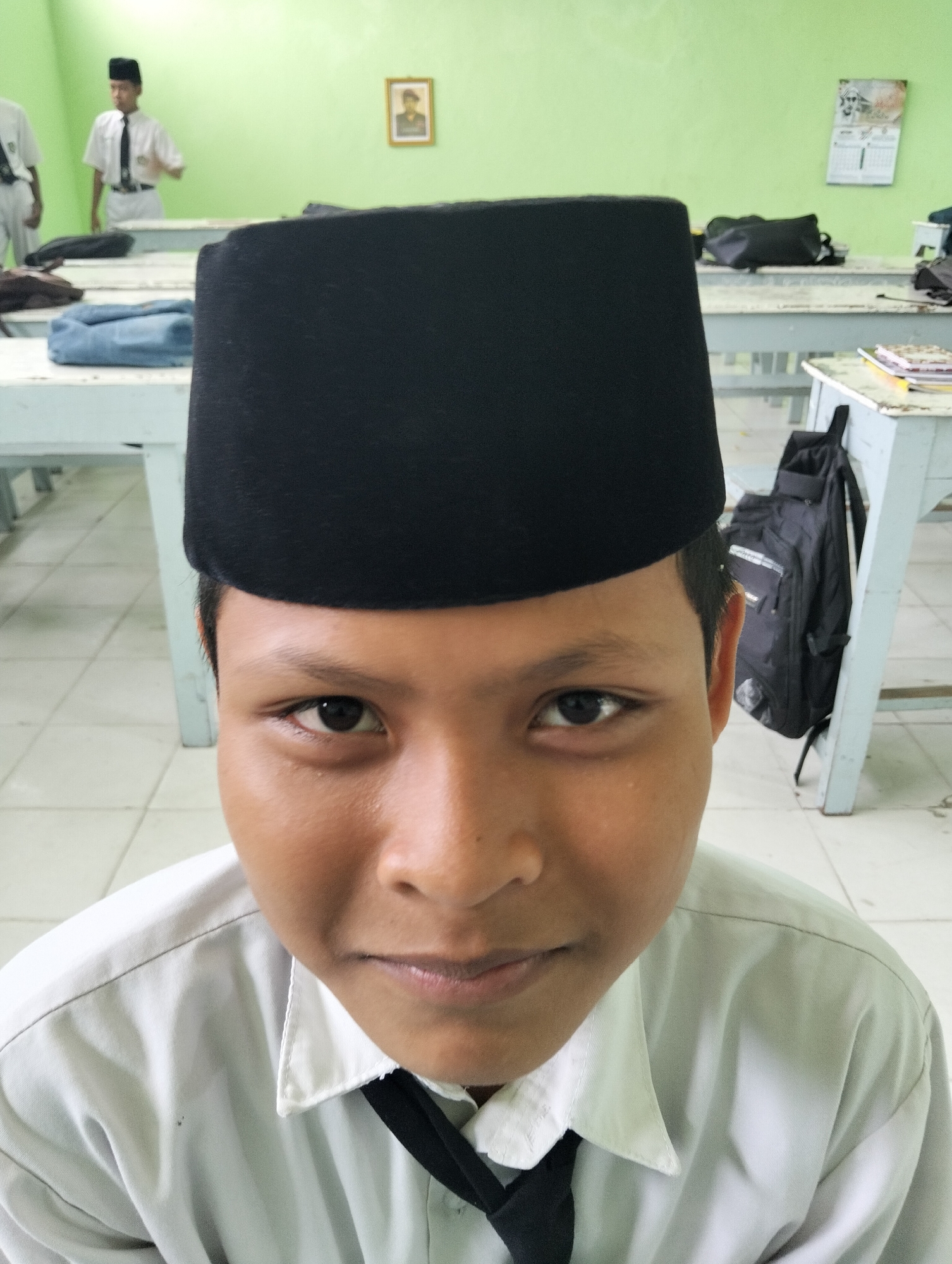 A young male, wearing a white collared shirt, a black tie, and a black peci (a traditional cap), is the central subject of this close-up, eye-level photograph. The background suggests a classroom setting. To the left, another male, also wearing a white shirt and black peci, is visible from the chest up. Desks are arranged in the room, with bags and books on them. The walls are painted green, and a framed picture hangs on the right wall. The floor is tiled. The lighting is bright, indicating daytime.