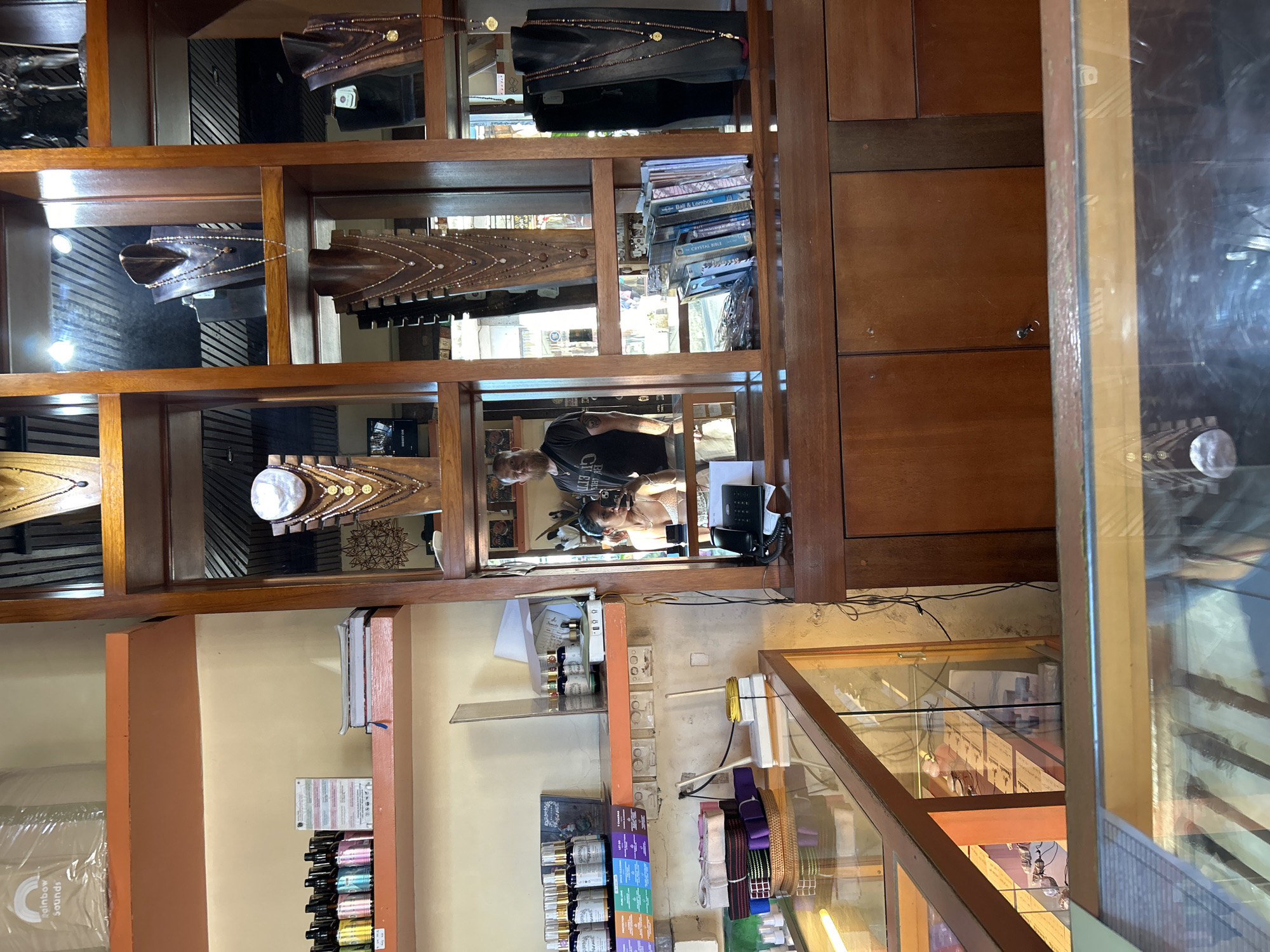 The image depicts the interior of a jewelry store in Ubud District, Indonesia. In the foreground, a glass display case is visible, containing various items that are not clearly discernible. Behind the display case, a wooden counter with a black landline telephone and a small white electrical device is present. To the right, a large wooden cabinet with multiple drawers and doors occupies the space.
In the background, a wooden shelving unit displays an assortment of jewelry, primarily necklaces on mannequins. The shelves are organized into compartments, with mirrors reflecting the items and the store's interior. On the top shelf, to the left, a rolled-up item wrapped in plastic with a "Rainbow Sounds" logo is seen. Below this, a shelf holds several bottles of what appear to be essential oils or perfumes, along with a stack of books.
The central focus of the background is a mirrored section that reflects two individuals. A man with a beard, wearing a black t-shirt, is standing on the left side of the reflection. A woman with dark hair, wearing a light-colored top, is on the right side of the reflection, looking into a small rectangular object, possibly a phone. They appear to be interacting within the store. The store's lighting is visible, with spotlights illuminating the jewelry displays. The overall environment suggests a retail setting, likely a boutique specializing in jewelry and possibly wellness products, consistent with the offerings found in Ubud.