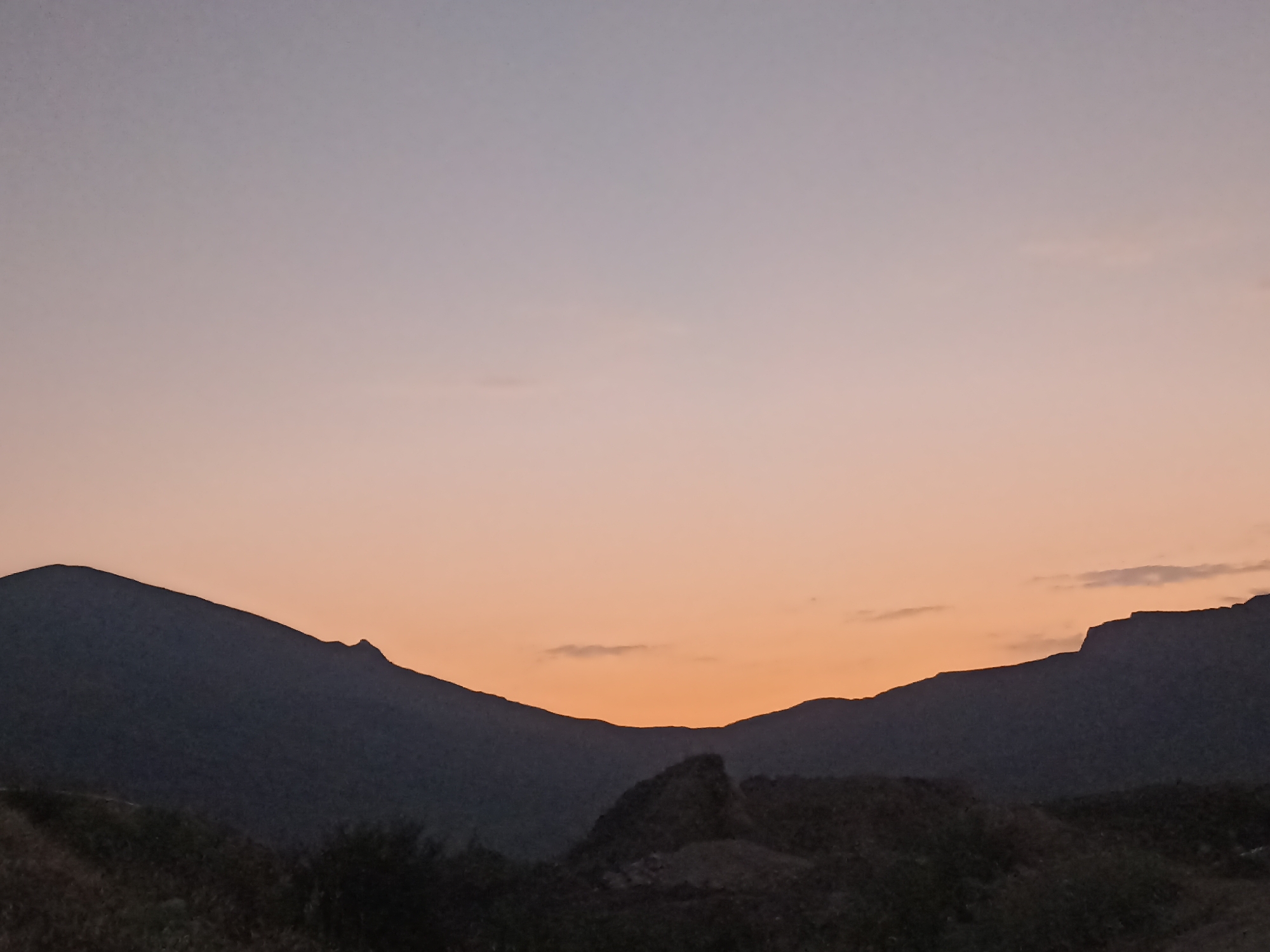 A silhouette of mountains against a gradient sky at dusk. The sky transitions from a pale purple at the top to a warm orange near the horizon. The mountains are dark, with jagged peaks and slopes. In the foreground, the dark shapes of vegetation and rocks are visible. The image appears to be taken from a natural landscape, possibly a mountainous area.