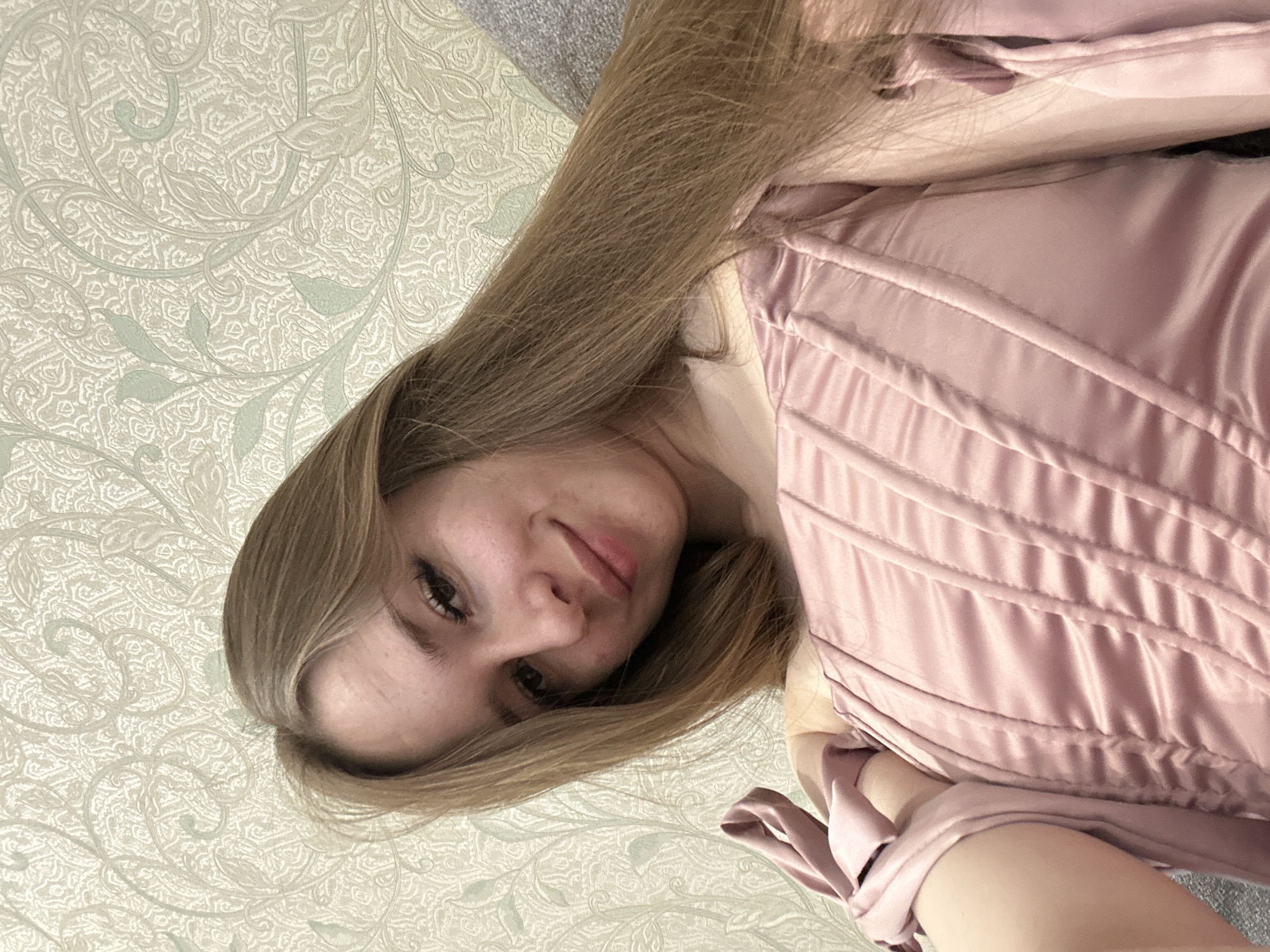 A woman is lying down, with her head on a patterned surface. She has long, light brown hair and is wearing a pink corset-like top with ribbon ties. The background is a patterned wall. The location is Unknown, Unknown.