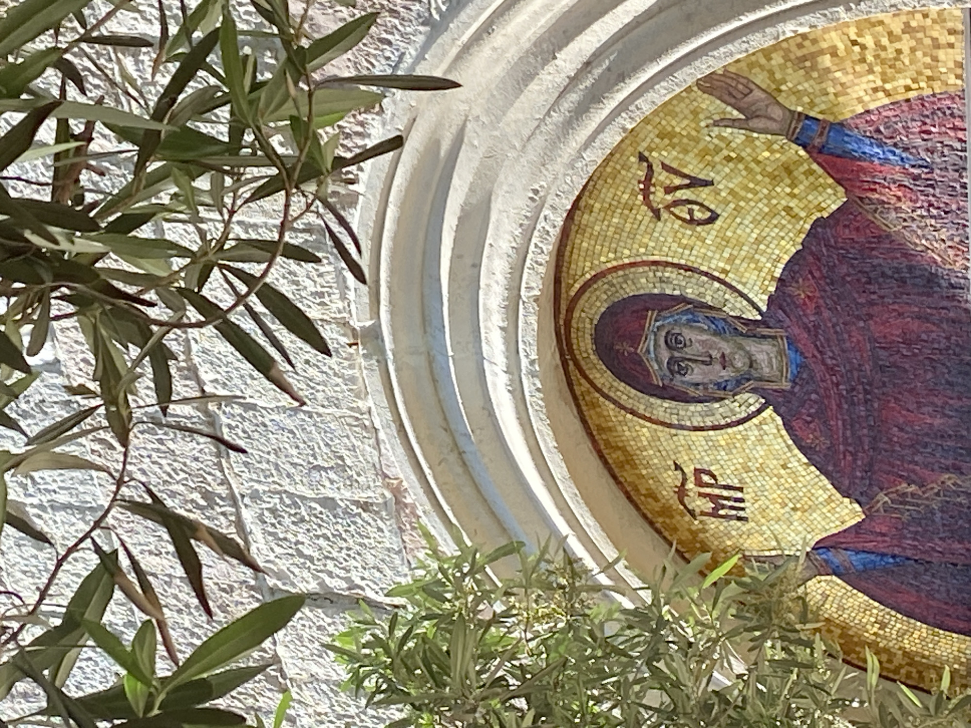 This image, captured in Budva, Montenegro, presents a detailed religious mosaic integrated into a light-colored stone wall, partially framed by vibrant olive branches. The mosaic vividly portrays a figure, likely the Virgin Mary, distinguished by a golden halo and draped in rich red and blue garments. Her solemn, forward-gazing face is depicted with thoughtful eyes, and her right hand is raised in what appears to be a traditional gesture of blessing or exhortation. The artistry of the mosaic is evident in its construction from countless small, brightly colored tesserae, with a dominant golden background highlighting the central figure.
The mosaic occupies an arched, deeply carved stone niche that forms part of a larger edifice constructed from textured, light-colored stones, suggesting historical or traditional architecture. The scene is bathed in bright sunlight, creating sharp shadows within the archway and indicating a clear day, likely during the brighter parts of the day. Lush green olive branches, characterized by their slender, elongated leaves, gracefully extend into the foreground from both the left and bottom right, gently softening the architectural lines and lending a natural, organic feel to the composition.
Inscriptions of Greek text are discernible on the mosaic's golden field. Above the figure's left shoulder, the letters "ΜΘ" (Mu, Theta) are visible, and below her right shoulder, "ΘΥ" (Theta, Upsilon) can be seen. These characters are traditional abbreviations for "Μήτηρ Θεοῦ" (Mētēr Theou), translating to "Mother of God," which affirms the identity of the depicted religious figure. The image conveys a serene and reverent atmosphere, blending cultural heritage with natural beauty typical of the Mediterranean region.
