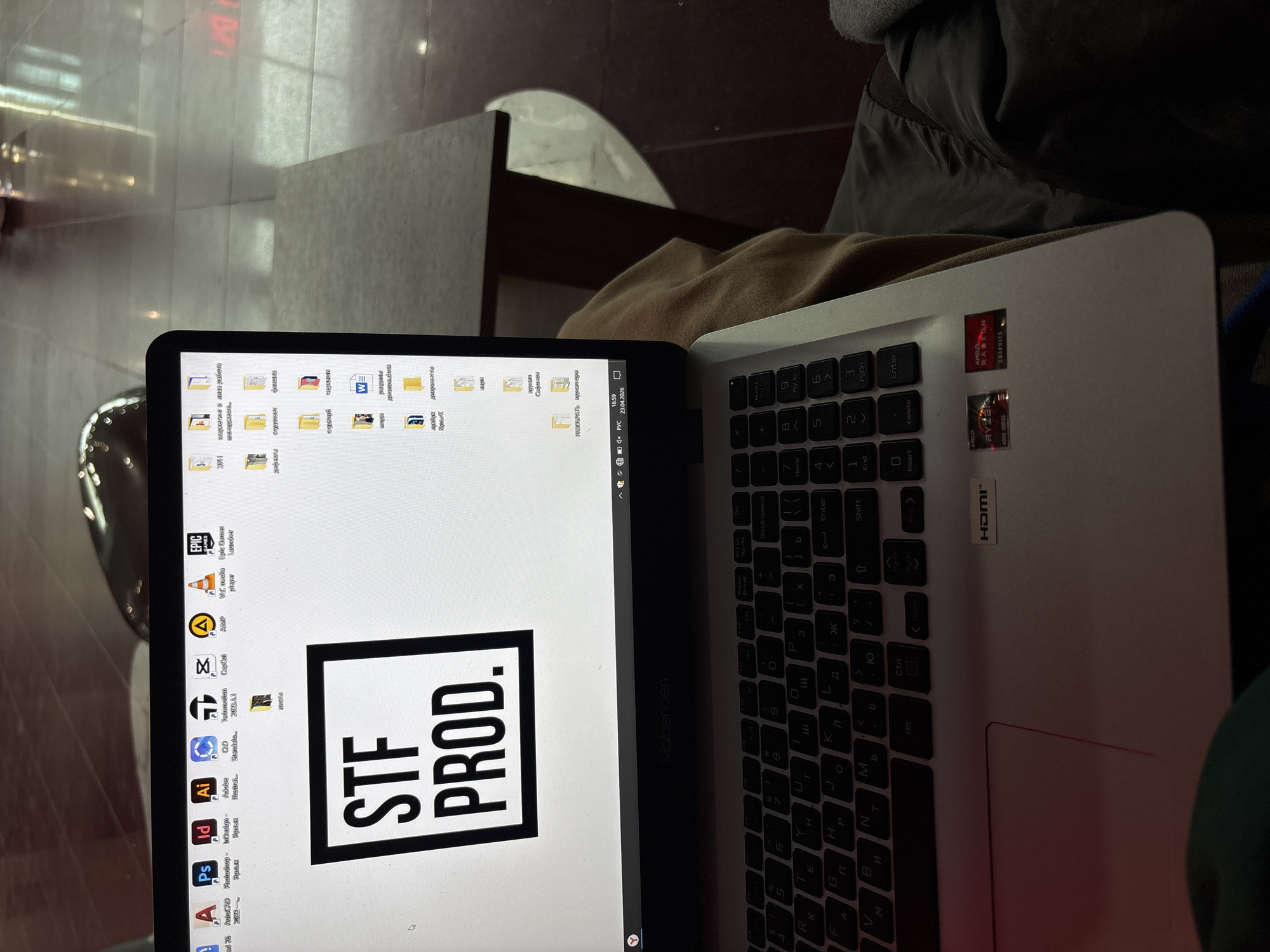 The image shows a laptop computer with a black screen displaying a logo that reads "STF PROD." and several application icons. The laptop keyboard is mostly black with white lettering; some keys have Cyrillic characters. A white laptop body has two stickers on the top right corner: One is a small red sticker, and the other one below has "RYZEN 4000 SERIES" with an "AMD RADEON GRAPHICS" red sticker. A silver "HDMI" logo sticker is also on the bottom. The open laptop is on someone's lap, covered with tan and gray clothing.
The screen desktop of the laptop indicates Russian folders. Icons include "AutoCAD 2022," "Photoshop," "InDesign," "Illustrator," "C4D," "Twinmotion," "CapCut," "AIMP," "VLC media player," and "Epic Games Launcher."
The photograph shows a blurred background of a public space with a dark chair. The floor background could be located in the city of Perm, Russia, indicated by the Cyrillic letters visible on the keyboard.