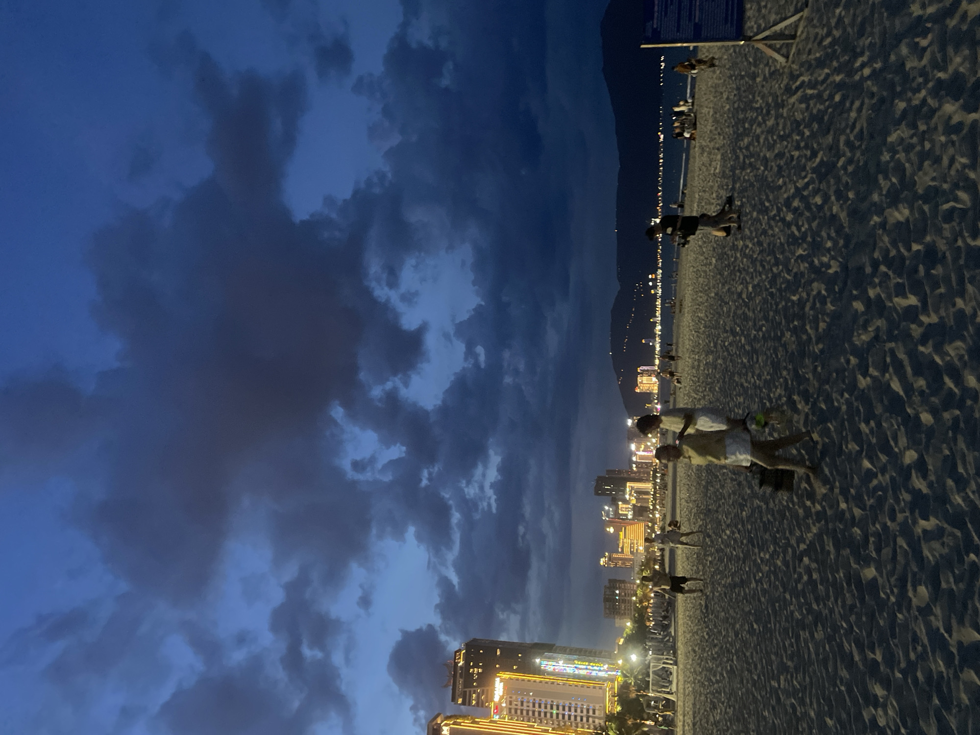 The image depicts a beach scene at dusk in the city of Da Nang, Vietnam. The sky is a deep blue with dark, dramatic clouds. In the background, a city skyline is illuminated with numerous lights, featuring tall buildings, including a prominent one on the left with colorful vertical lighting. The beach itself is covered in sand, with visible footprints and ripples. Several people are scattered across the sand, some walking, some appearing to be engaged in activities. In the mid-ground, a woman in a yellow top and white skirt is walking towards the right, carrying a bag. To her left, two other individuals are walking together. Further to the right, a man in a black shirt and shorts is walking with a dog. The ocean is visible in the distance, with a line of lights indicating the coastline. The overall atmosphere is that of a lively evening on a coastal city beach.