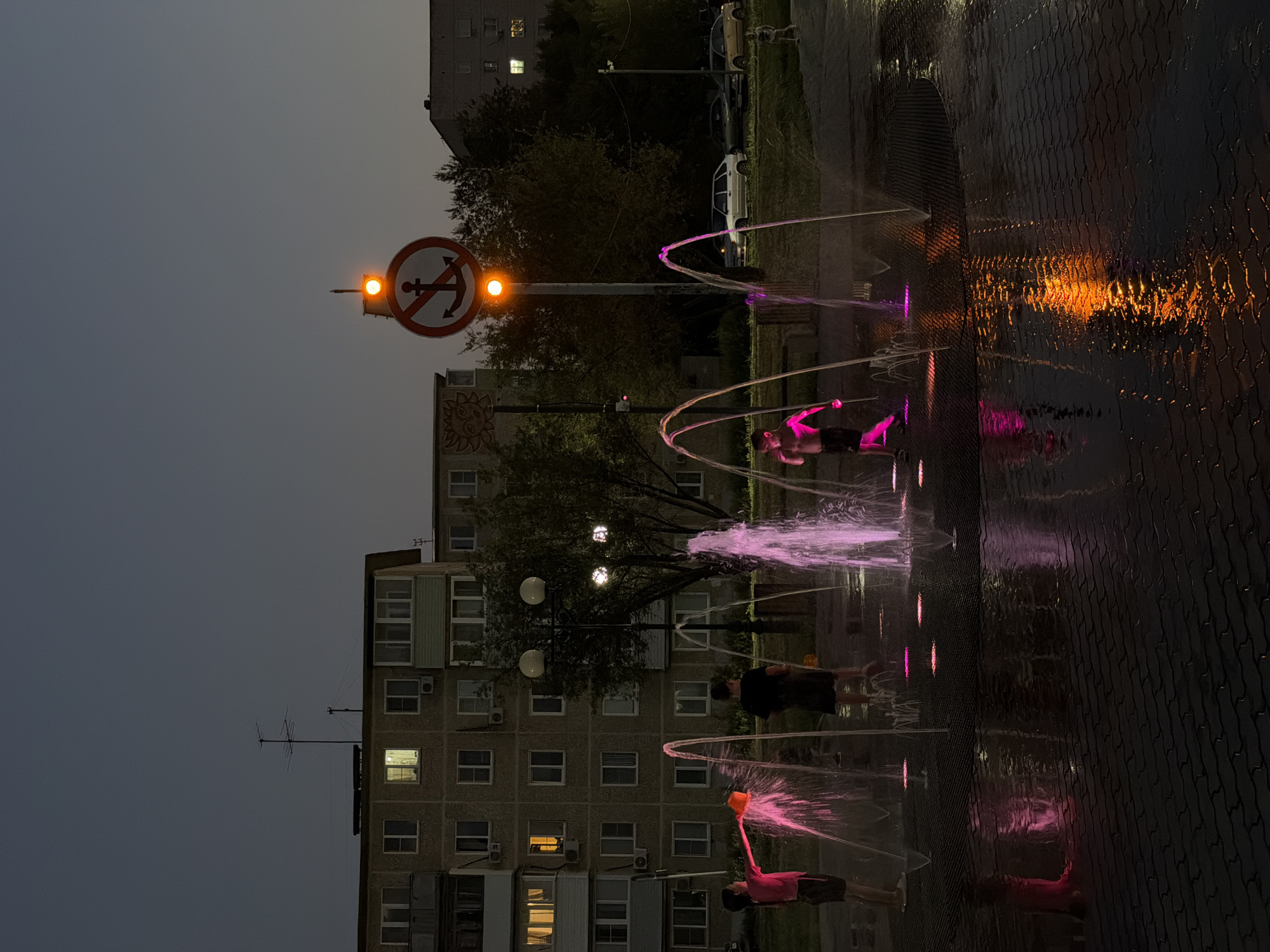 At dusk in Kamyshin, Russia, a splash pad is active with children playing in the illuminated water jets. Several young people are visible, one girl in a pink shirt is holding an orange bucket, splashing water. Another child, shirtless, stands amidst the spray, illuminated by pink and purple lights. The splash pad is set on a dark, patterned pavement, which is wet and reflects the colorful lights. In the background, a multi-story apartment building with visible windows, some lit, dominates the scene. A street lamp with two spherical lights stands near a tree. Further in the background, cars are parked, and a traffic signal with an illuminated orange light and a "no anchoring" sign is mounted on a pole. The sky is a dark, dusky blue.