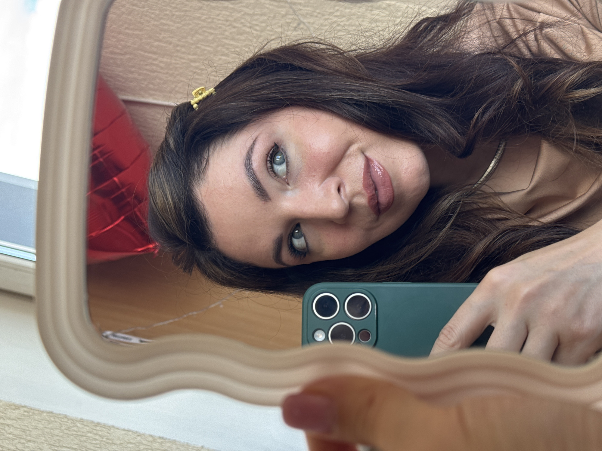 The image shows a woman taking a selfie in a mirror. The mirror has a wavy, beige frame. The woman has brown hair with a gold hair clip, light eyes, and is wearing a light brown top and a silver necklace. She is holding a green smartphone with multiple camera lenses. A red balloon is partially visible in the mirror behind her. The background shows a window and a light-colored surface. The location in the city of Kryvyi Rih, Ukraine, is not discernible from the image.