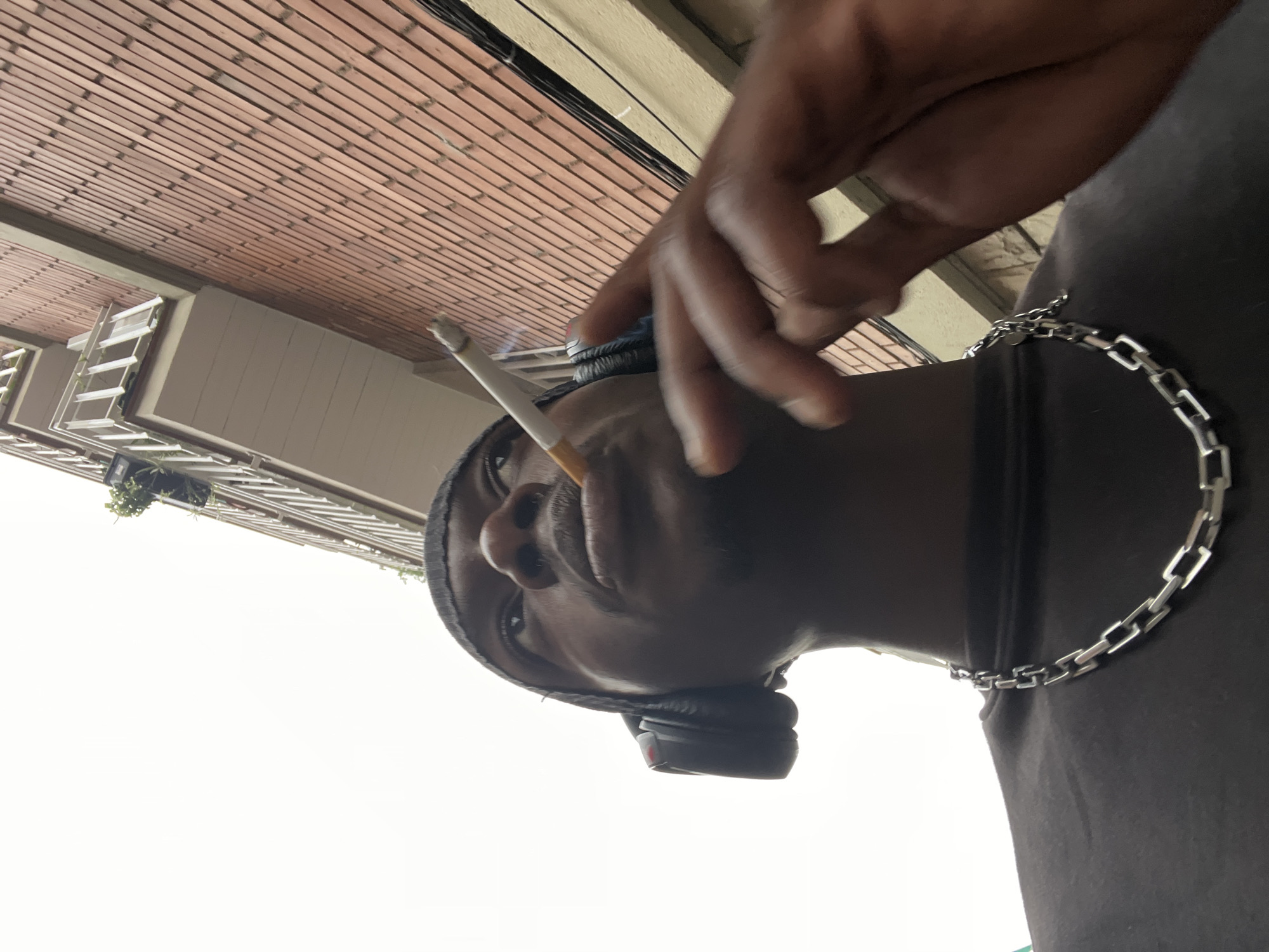 A low-angle, portrait-oriented photograph depicts a dark-skinned man with a cigarette in his mouth. He is wearing a silver chain necklace and headphones. A building with a brick exterior and a balcony with plants is visible in the background. The setting appears to be an urban area, possibly Barcelona, Spain, based on the architectural style. The sky is visible in the background, suggesting an outdoor setting.