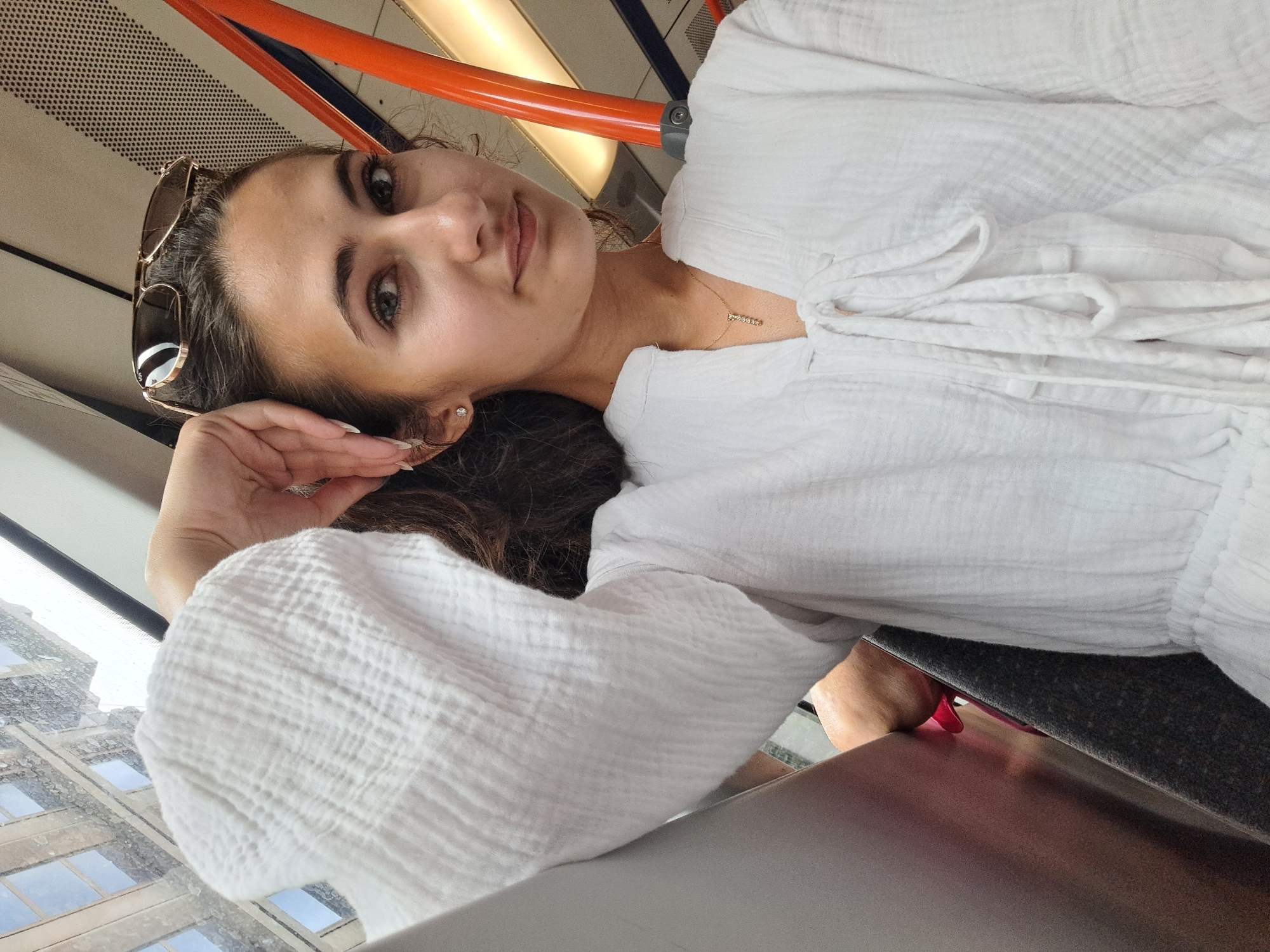 A young woman with long, dark hair and brown eyes is captured in a close-up, selfie-style shot. She is wearing a white, crinkled fabric top with puffed sleeves and a delicate gold necklace with small charms. Sunglasses are perched on her head. She is resting her head on her hand, and her expression is relaxed and serene.
The background suggests an indoor setting, possibly a mode of transport, with an orange handrail and a textured ceiling visible. A window to her left reveals a glimpse of city buildings, hinting at an urban environment. The lighting appears to be natural, suggesting daytime. The image is oriented sideways. There is no visible text.
