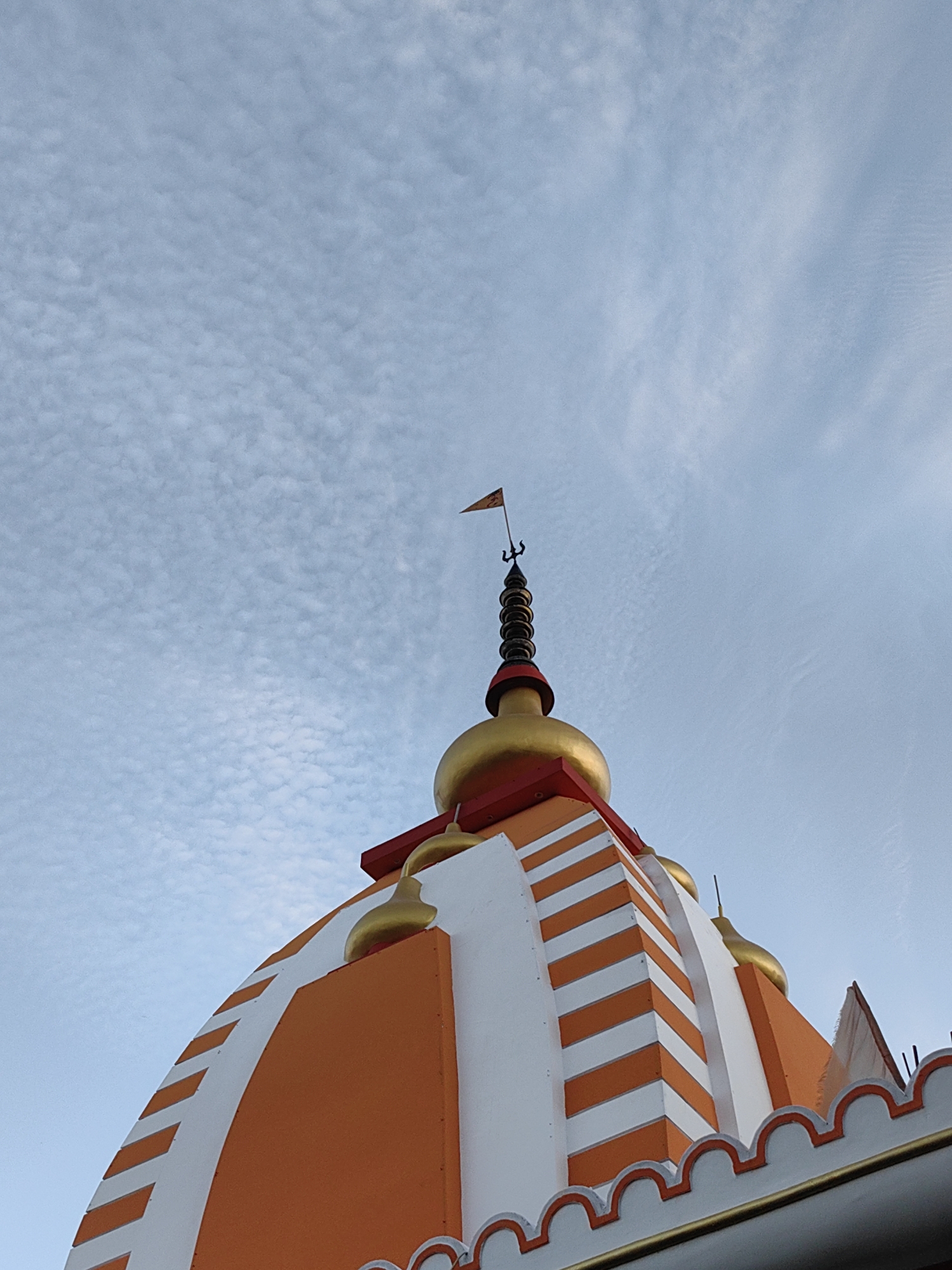 The image shows the top of an orange and white striped dome of a temple, against a cloudy blue sky. A golden spire with a flag and trident adorns the very top of the dome. The temple architecture appears to be of Indian origin. The photograph was taken in Sonauli, India.
