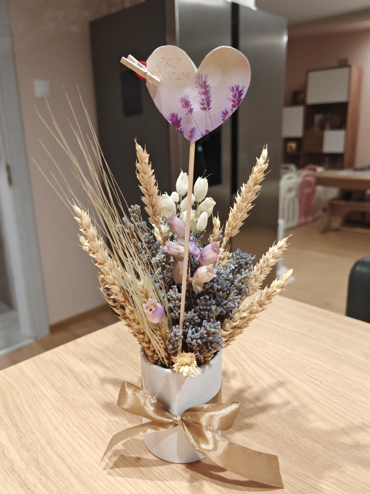 A close-up shot captures a decorative arrangement of dried flowers and wheat stalks in a small white ceramic pot, placed on a light-colored wooden table. A shimmering gold ribbon is tied around the neck of the pot. Atop a wooden skewer, a heart-shaped cutout adorned with lavender sprigs and the inscription "C&C 01.07.25" is pinned with a tiny clothespin. The background is blurred, showing a large, dark kitchen appliance, shelves with items, and a glimpse of a dining area with furniture. There are no people or animals visible in the image, and the scene is static. This image was taken in Plovdiv, Bulgaria.