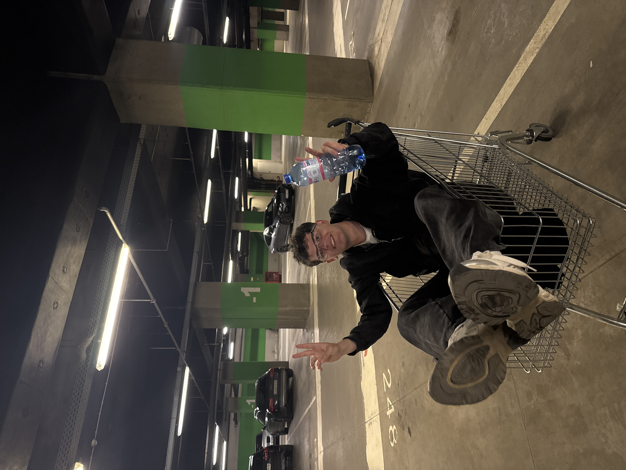 The image shows a young man with glasses inside a shopping cart in an indoor parking garage. He is holding a bottle of water. The pillars of the parking garage are marked with green paint at the top. There are cars parked in the background, indicating parking spaces. The floor is marked with parking space numbers, one of which reads "248". The image is rotated at an angle. The location is possibly in Sofia, Bulgaria.