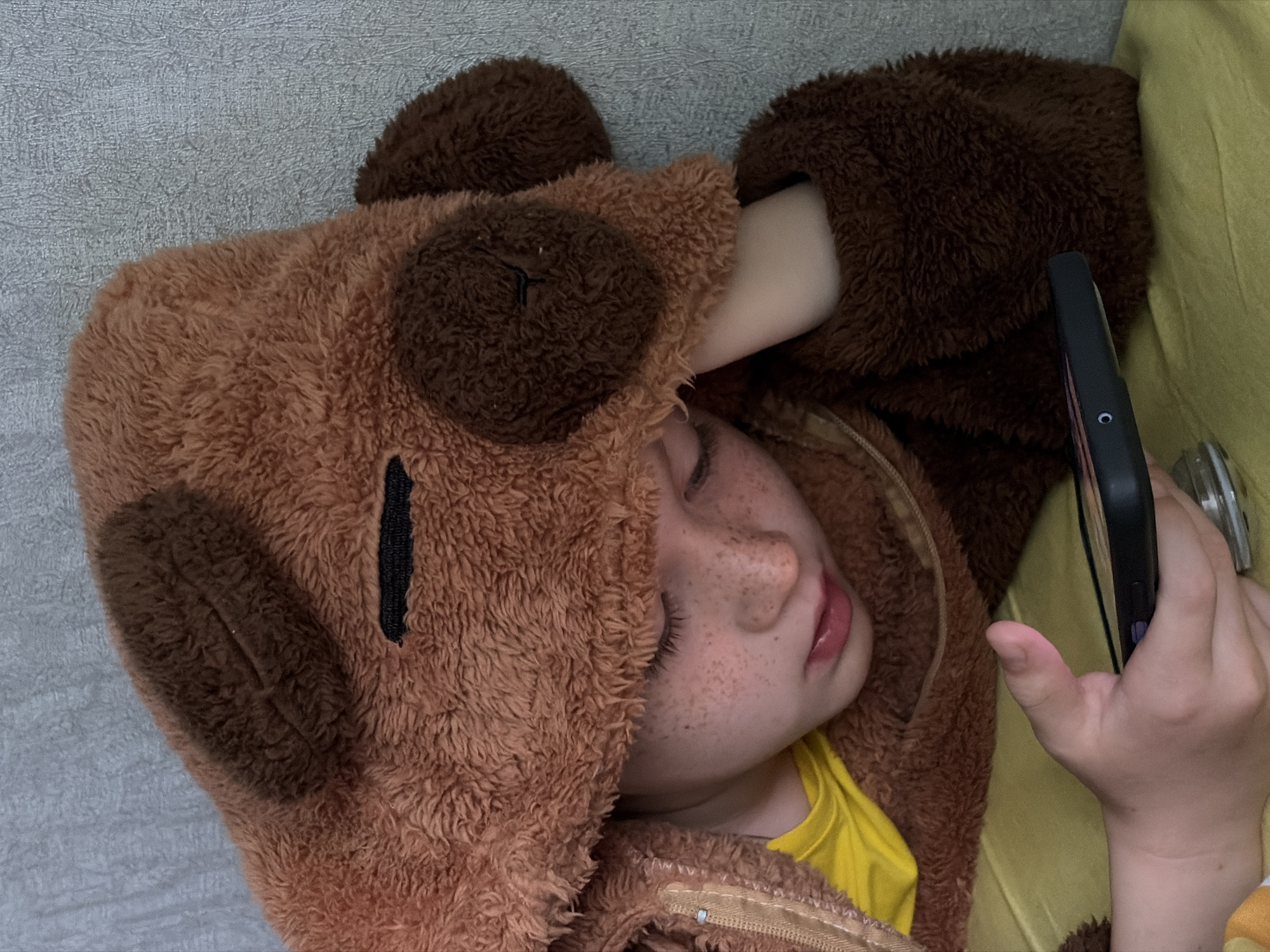 A young child, likely of preschool or early elementary age, is depicted resting indoors in Chaikovsky, Russia. The child appears to be asleep or deeply relaxed, lying on a textured grey surface, possibly a sofa or bed. They are wearing a cozy, fluffy brown hooded outfit, resembling an animal like a bear or dog, complete with darker brown patches on the hood for ears or facial features, and a vertical black strip near the center of the hood. A bright yellow t-shirt is visible beneath the brown garment, which also features a visible zipper.
The child's face, partially framed by the hood, shows closed eyes and a slightly open mouth. Their nose and cheeks are covered in numerous light-colored freckles. In their right hand, they are holding a black smartphone enclosed in a case, with a small white circular detail visible on its side. A section of plain yellow or light green fabric is visible in the background, to the right of the child. The image conveys a sense of peaceful rest. No visible text is present in the image.