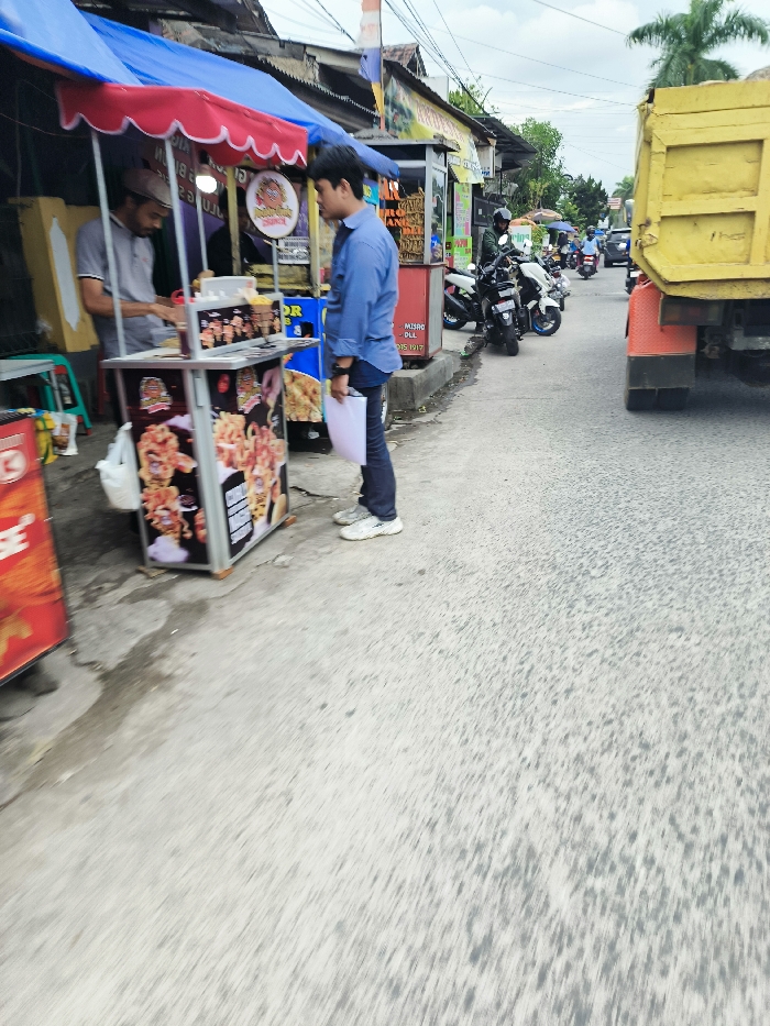 The image shows a street scene in Kecamatan Dayeuhkolot, Indonesia, located at latitude -6.963 and longitude 107.595. A man in a blue shirt and white sneakers is standing in front of a food stall, looking at a food item in a container. Another man in a light-colored shirt and black pants is standing behind the stall, looking towards the man in blue. The stall is made of metal and has a blue and red awning, displaying a picture of food. The street is paved and has a light gray color. Several motorcycles are parked on the side of the street, and a yellow truck is driving away. There are some buildings in the background, all painted with a variety of colors. The sky is cloudy.