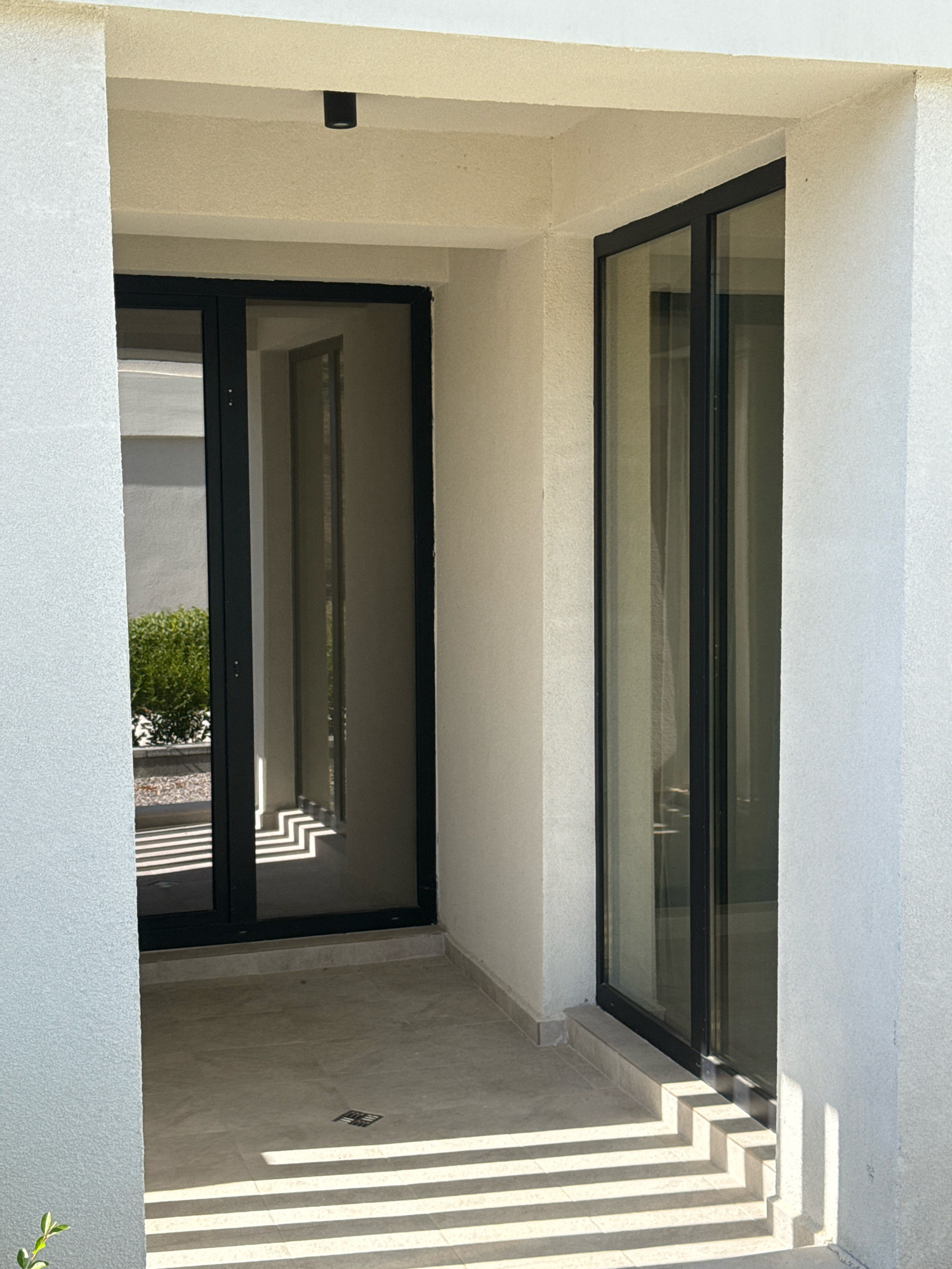 The image shows a modern outdoor patio with large glass sliding doors. The patio is paved with light-colored tiles, and there are strong shadows cast across the floor, indicating sunlight. To the left, a grey textured wall of a building is visible, and to the right, a white textured wall. The sliding doors are framed in black. Through the left door, a glimpse of green bushes and gravel can be seen. A black cylindrical light fixture is mounted on the ceiling of the overhang above the doors. The scene appears to be located in Sozopol, Bulgaria.