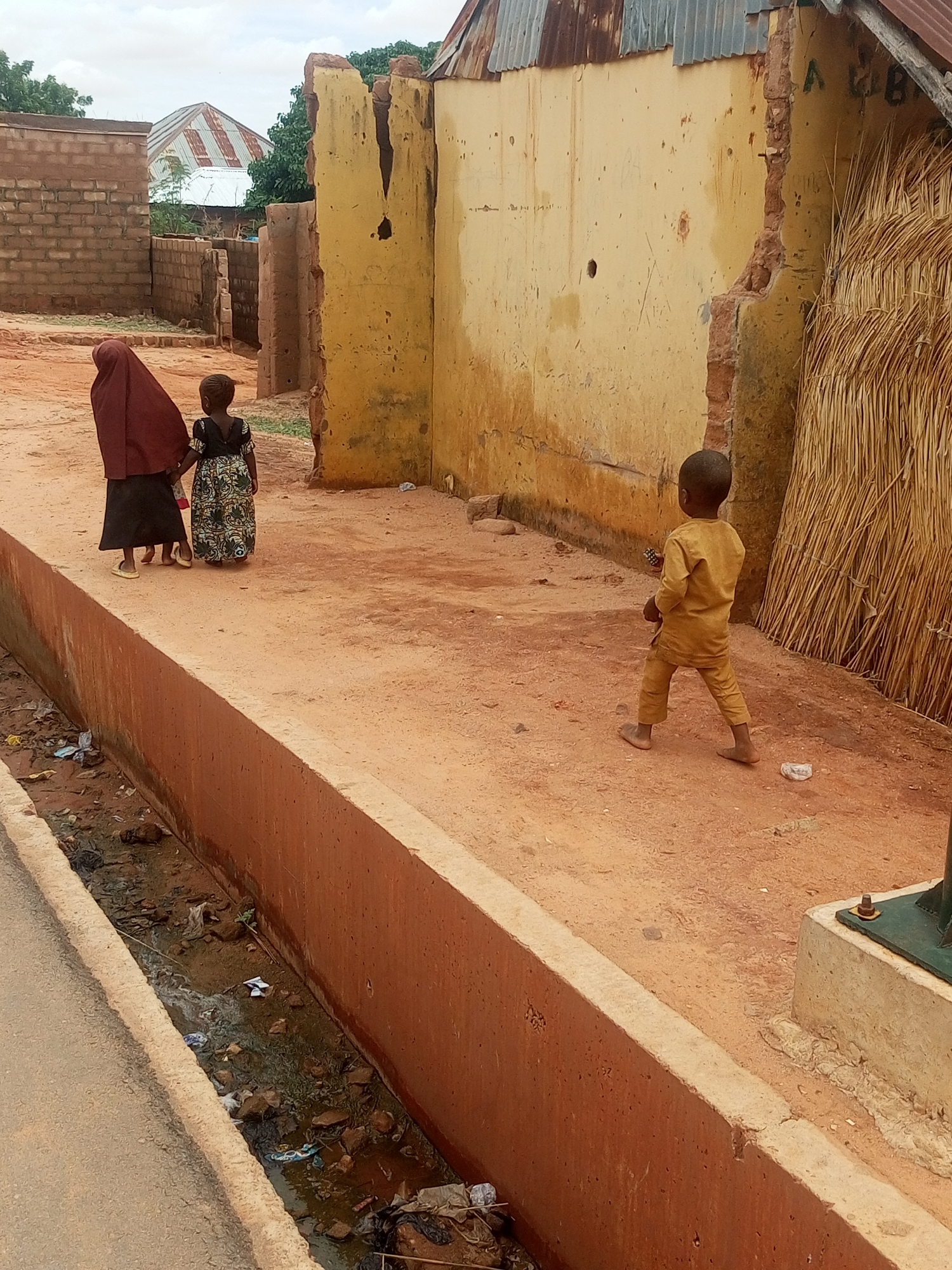 The image depicts an outdoor scene in the city of Unknown, Unknown, featuring three individuals and several structures under an overcast sky. The ground throughout the visible area consists of reddish-brown packed dirt.
On the left side, an open concrete drainage ditch, approximately 60-70 cm wide and 40-50 cm deep, runs parallel to the viewer. This ditch contains stagnant, murky water mixed with various pieces of refuse, including plastic bags, paper, and other debris.
In the mid-ground, slightly left of center, an individual clad in a dark red head covering and a dark dress walks away from the camera, holding the hand of a smaller child. This child is wearing a dress with a dark upper portion and a patterned skirt featuring green, white, and black designs. Both individuals are walking towards the background.
To the right of the central axis, a young child, likely a toddler, wearing a light tan or yellowish long-sleeved shirt and matching pants, is walking away from the camera. This child is barefoot and holds a small, dark, rectangular object, possibly a toy or remote control, in their right hand.
The primary background structure on the right is a building with faded yellow plaster over what appears to be reddish-brown mud brick or unplastered brick. This wall exhibits significant structural damage, including large vertical cracks and sections where the plaster has fallen away, exposing the underlying material. On the far right, against this yellow wall, a large stack of dried, light-brown fibrous material, possibly straw or reeds, is leaning vertically. Above this, a partially visible, weathered corrugated metal roof is present, and faint dark lettering, possibly "A B", can be discerned on the upper part of the yellow wall.
Further in the background, to the left, are additional buildings. One has exposed reddish-brown brick walls, and behind it, another structure features a rusted corrugated metal roof against the pale sky. Adjacent to the ditch on the foreground right, a concrete base with a metal bolt is partially visible, suggesting a foundation for a pole or similar fixture.