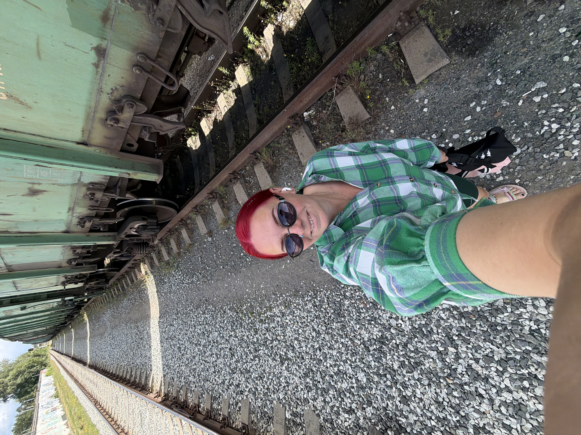 This media file shows a person with vibrant red hair, wearing sunglasses and a green and white plaid shirt. They are standing on a gravel embankment next to a train track, with a green train car visible to their left. The person is smiling and appears to be taking a selfie, as their arm holding the camera is visible in the foreground. A black bag with pink accents and a pair of sandals are on the ground near the person. The scene suggests an outdoor setting, likely during the daytime, with clear weather. The location is identified as Chaikovsky, Russia. The overall mood seems relaxed and casual. No significant text is visible in the image, apart from some faint markings on the train car.