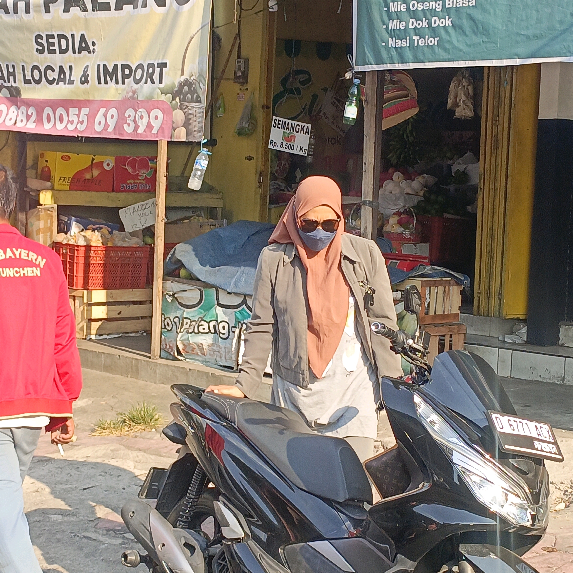 The image captures a street scene in Salatiga, Indonesia, outside a market stall. A person wearing a pink headscarf and sunglasses, with a gray jacket over a light-colored shirt, stands next to a black motorcycle. The motorcycle has a license plate that reads "D 6771 ACX". In the background, a stall displays various items, including fruit, produce, and bottled drinks. Signs advertising "SEMA NGKA Rp. 8.500/Kg" (Watermelon Rp. 8,500/Kg) and a phone number "0882 0055 69 399" are visible. Another person, seen from behind and wearing a red jacket with "BAYERN MUNCHEN" printed on it, walks away from the stall, holding a cigarette. The environment appears to be an outdoor marketplace with sunlight casting shadows on the ground.