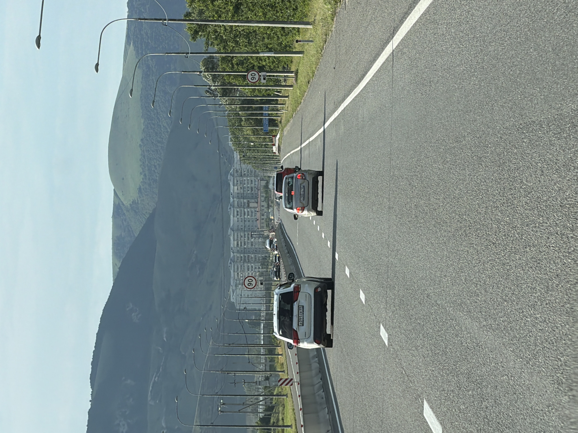 The image shows a view from a moving vehicle on a highway. The highway is paved and has white lane markings. On the left side of the highway, there is a steep, tree-covered mountain range. A row of streetlights lines the side of the road, curving upwards towards the mountain. In the distance, there are buildings that appear to be part of a city. Several cars are visible on the highway, all traveling in the same direction. One car, a white SUV, is in the foreground, and another car, a red hatchback, is behind it. There is a speed limit sign visible on the left side of the road, indicating "90". The sky is a light blue, suggesting it is daytime with clear weather. The scene appears to be set in a mountainous region with an urban area nearby. The license plate on the white SUV is visible and appears to read "A091CB 92". The location context is given as Kubina, Russia. There are no people clearly visible in the image. The overall impression is one of travel through a scenic and developed area.