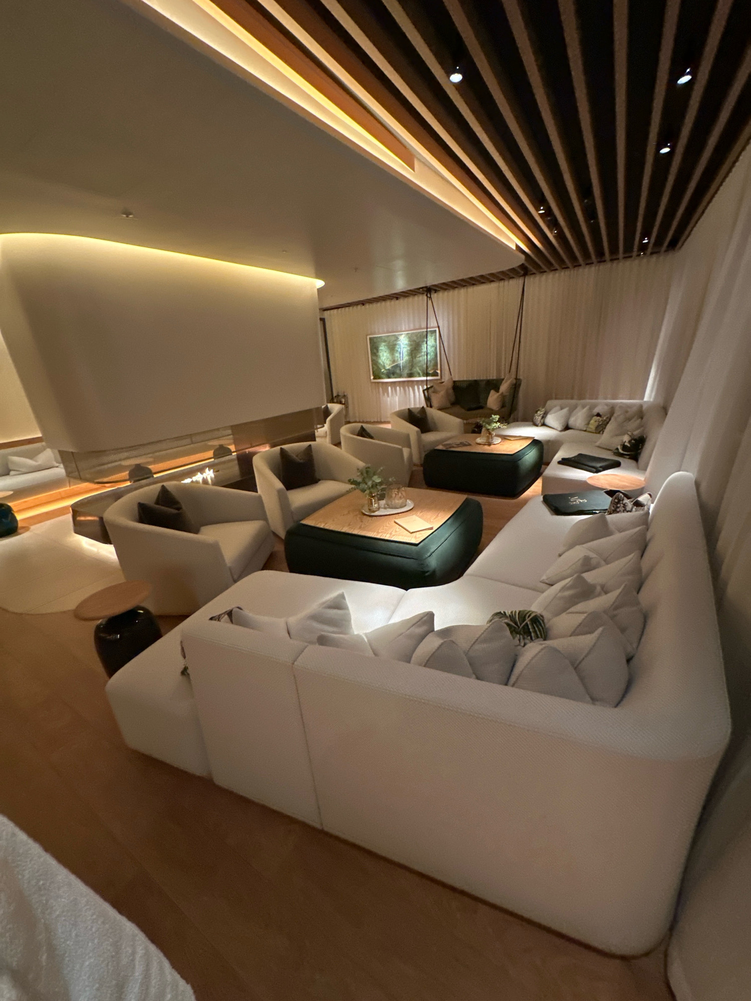 The image depicts a modern, luxuriously furnished lounge area. The space features a large, sectional white sofa with numerous plush pillows. In the center of the seating area are two dark green ottoman-style coffee tables with wooden tops, one displaying a small plant and another holding a book or menu. Around the perimeter, there are several cream-colored armchairs, some with dark throw pillows. A contemporary fireplace with a glass screen is visible in the background, with a subtle flame. The ceiling is a distinctive architectural feature with wooden beams and integrated lighting, creating a warm ambiance. Drapes of sheer white fabric hang in the background, adding a soft, ethereal quality to the room. A framed artwork depicting a forest scene hangs on the wall. The flooring is polished wood. The overall impression is one of a sophisticated and comfortable waiting or relaxation area, potentially in a high-end hotel, spa, or private club. The scene is devoid of people, suggesting a quiet or unoccupied moment. The lighting is warm and indirect, with recessed ceiling lights and under-lighting along architectural features. No specific location in London, United Kingdom is identifiable from the visual elements present.