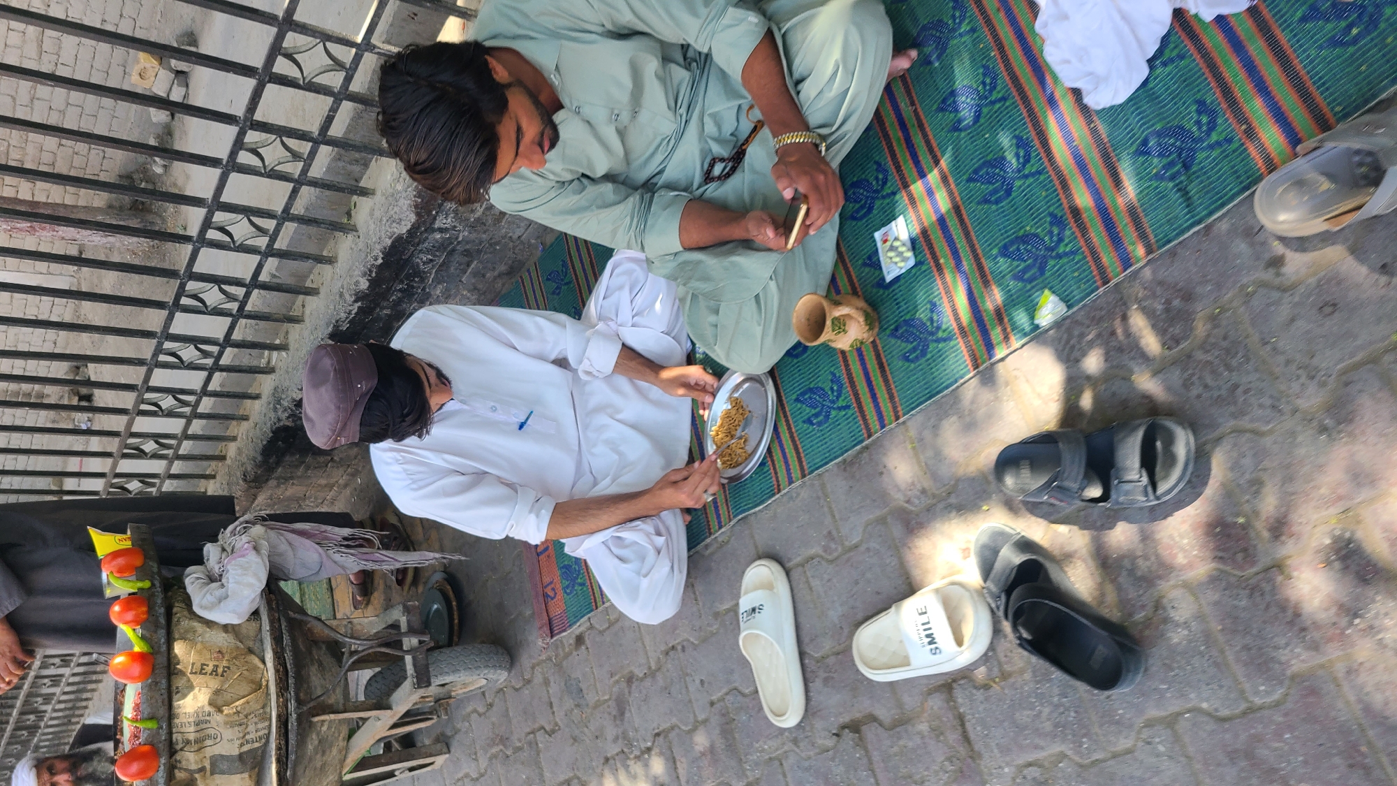 Two men are sitting on a patterned mat on a paved surface, located near a metal fence. One man is wearing a light green outfit and holding a phone. The other man is wearing a white outfit and a brown hat, holding a plate with food. A clay pot sits between them. Nearby are various pairs of sandals. A food cart with tomatoes and other items is visible in the background. The scene appears to be outdoors, possibly in a public area in Quetta City Tehsil, Pakistan.
