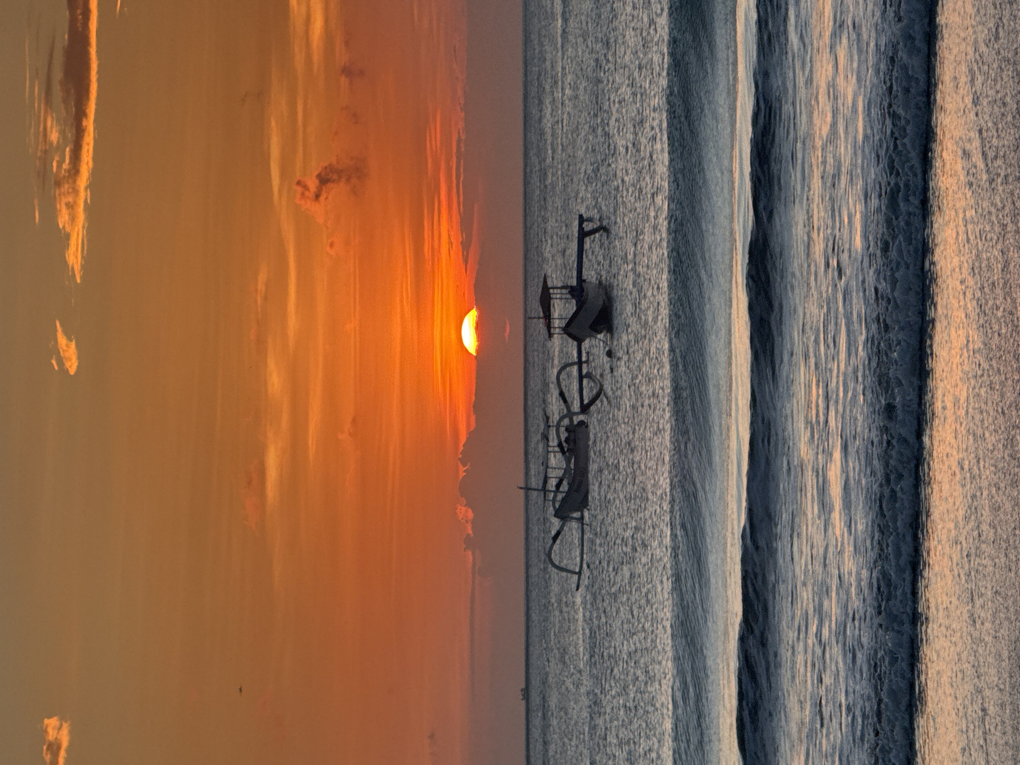 The image captures a sunset in Kuta Selatan, Indonesia, as seen from the beach. The setting sun, partially obscured by clouds, casts an orange and yellow glow across the sky. The ocean reflects this light, creating a textured pattern on the water's surface. Two traditional outrigger boats are moored in the water, silhouetted against the sun. The beach is not visible, with the view focused on the water and sky.