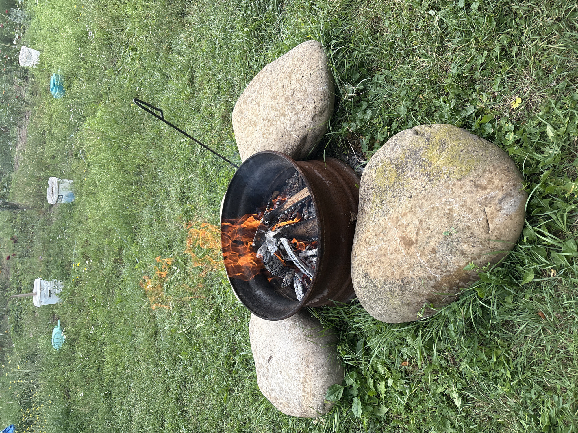 A campfire burns in a metal fire pit on a grassy lawn. Three large, smooth stones are arranged around the fire pit. In the background, several white buckets and a blue watering can are scattered across the lawn. The scene appears to be in a backyard or outdoor recreational area in Petrykiv, Ukraine.