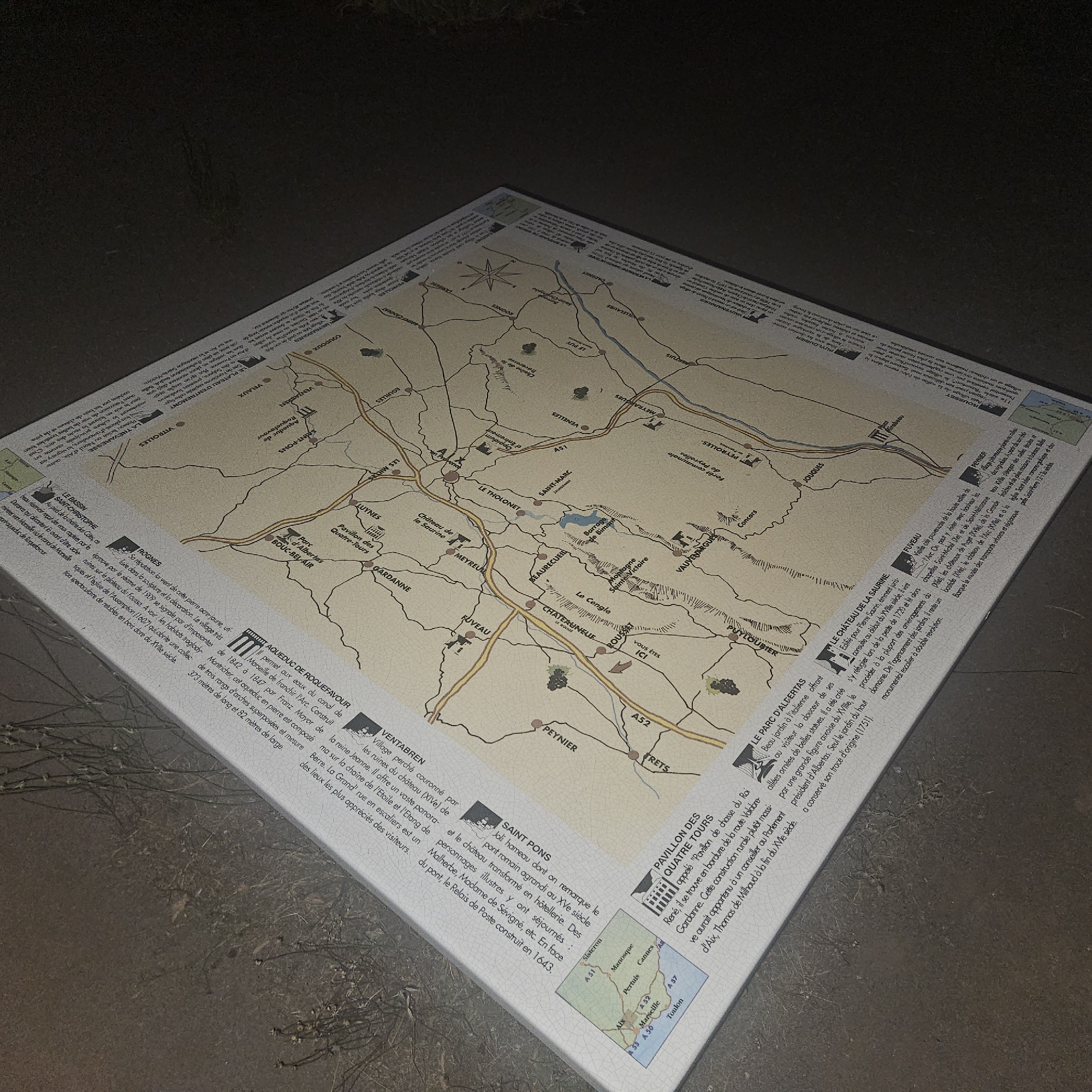 The image shows a map and informational panel laid out on a dark, textured surface, possibly the ground at night. The map displays a region with roads, towns, and geographical features. Various points of interest are labeled on the map with text in French, indicating historical sites, villages, and parks. The informational panel provides detailed descriptions of these locations, also in French. One of the highlighted locations is "ROUSSET," described as being where "VOUS ETES ICI" (You are here). Other prominent labels on the map include "AIX," "MARSEILLE," and "TOULON." The overall scene is dimly lit, with the map and panel illuminated by an unseen light source, creating strong shadows and highlighting the texture of the map's surface and the surrounding ground.