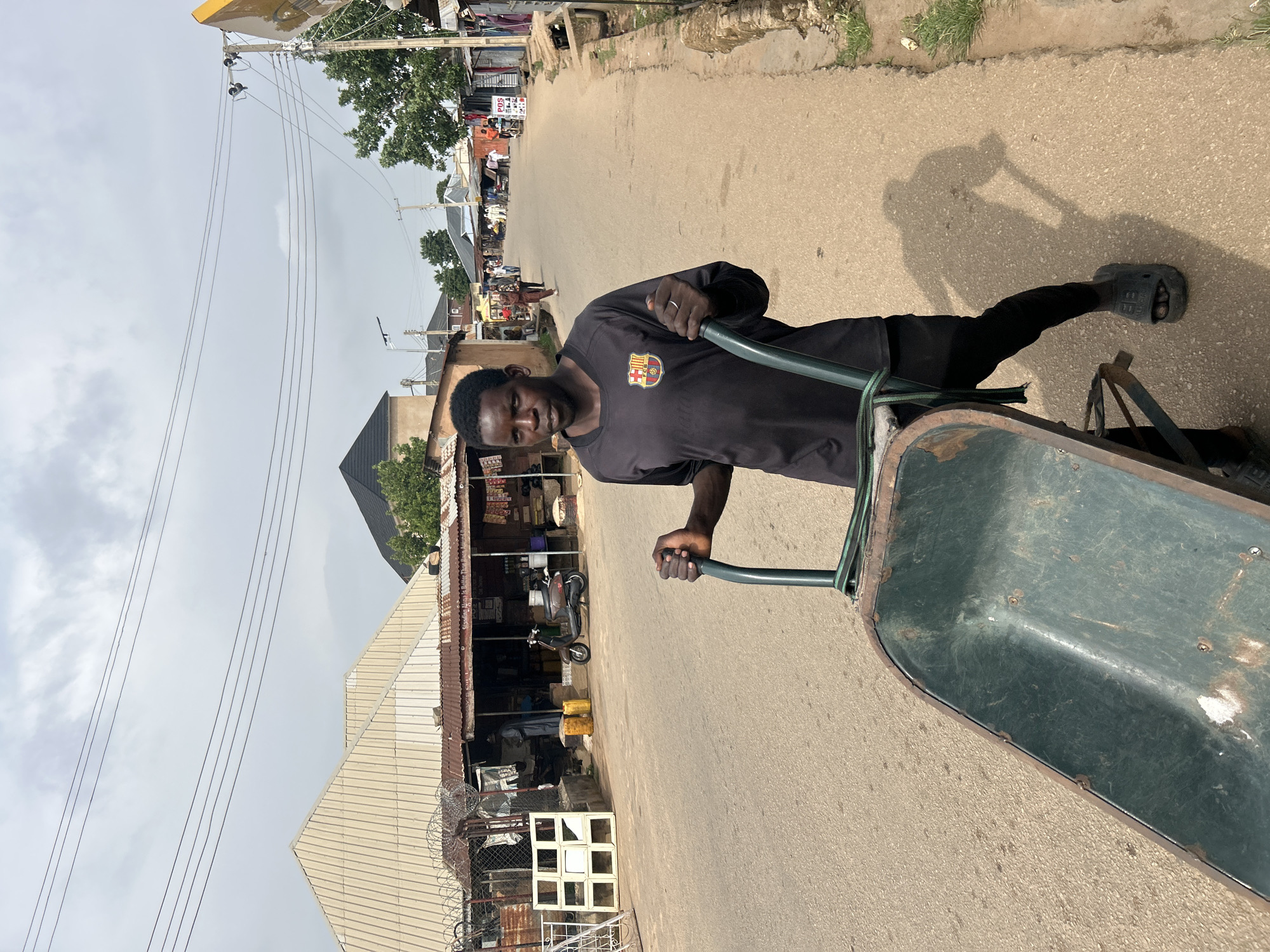 In Jos, Nigeria, a man is captured from the waist up, pushing a dark green wheelbarrow down a paved street. He is wearing a black long-sleeved shirt with a red and yellow emblem on the left chest and black pants. The wheelbarrow is positioned at an angle, with its handles prominently in the foreground. The street is lined with shops on both sides, many with corrugated metal roofs. In the background, people are visible walking, and a scooter is parked in front of one of the shops. The sky is overcast, with a network of overhead wires crossing the frame. The overall environment suggests a busy market or commercial area in the city.