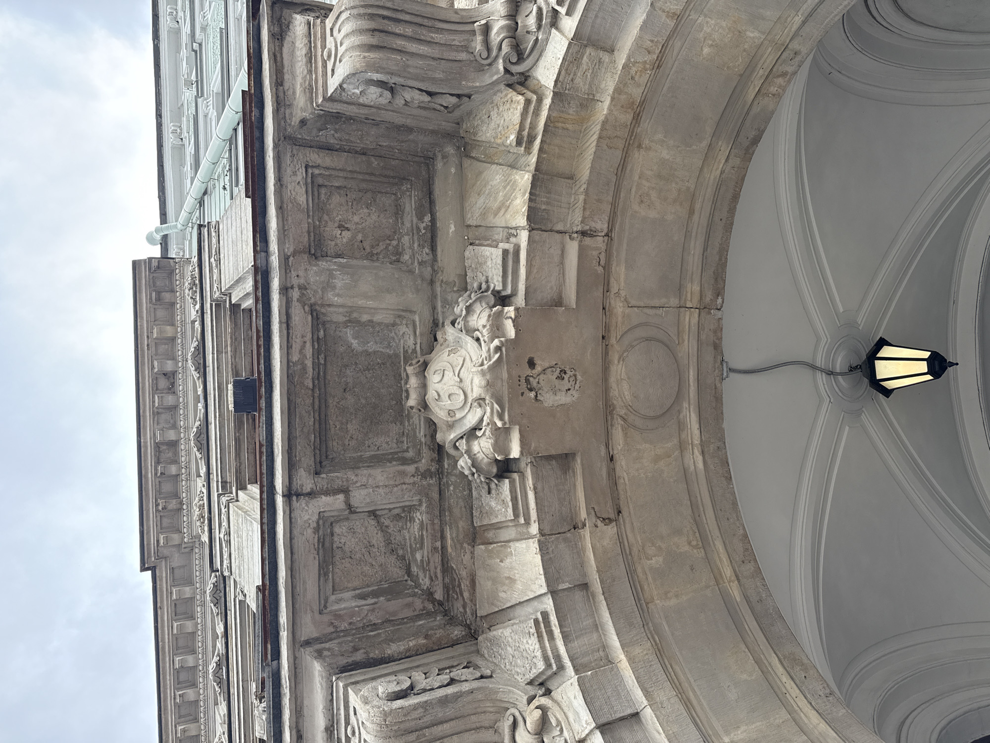 The image shows an architectural detail of an old building in Saint Petersburg, Russia, specifically an ornate stone archway with decorative elements. Above the arch, there is a section of facade with carved stone molding and a cartouche containing the numbers "62" and a coat of arms. Further up, a downspout is visible along the side of the building. The underside of the archway reveals a vaulted ceiling with a hanging lantern. The sky is overcast.