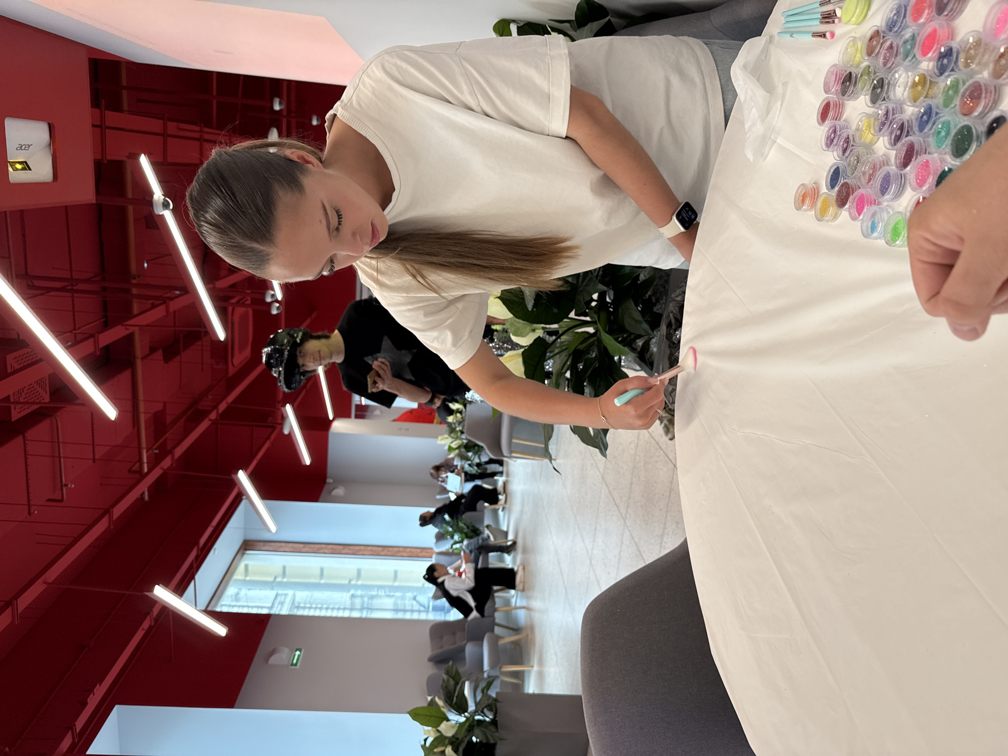 The image depicts an indoor scene in Vladivostok, Russia, featuring a creative workshop or activity in a modern, open-plan setting. The main subject is a young woman with light brown hair tied in a ponytail, wearing a white t-shirt and a white smartwatch. She is leaning over a table covered with a white sheet, holding a small brush with a pink tip, and appears to be applying glitter or pigment to the surface, demonstrating a focused expression.
On the table, to her right, a large collection of small, clear plastic containers filled with various colors of glitter or powdered pigments is neatly arranged. Several brushes with blue handles are also visible among the containers. Another person, partially visible behind the main subject, also appears to be participating in the activity.
The background reveals a distinctive and modern interior design. The ceiling and upper walls are painted a vibrant red, featuring numerous white, linear fluorescent light fixtures. An "acer" logo is visible on a white projector or device mounted on the red wall. Further back, several individuals are seated on lounge chairs in what appears to be a common area or waiting space, some engaged with laptops or phones. Potted green plants are placed around the room, adding natural elements to the contemporary environment. A large window in the far background suggests ample natural light, indicating it is daytime. The scene conveys a relaxed yet engaging atmosphere, likely a recreational or team-building event within a corporate or public venue.