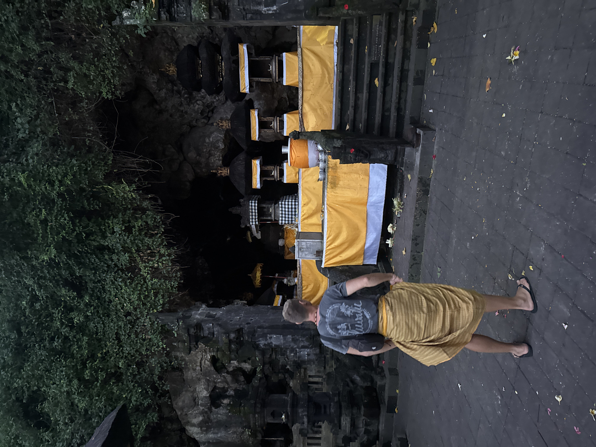 In the city of Dawan, Indonesia, a man stands facing a religious shrine in front of a cave entrance. He wears a grey t-shirt with a design, a yellow sarong wrapped around his waist, and flip-flops. A black crossbody bag is slung over his shoulder. The shrine is decorated with yellow fabric and features several tiered structures topped with dark, pointed roofs. Umbrellas with yellow fabric are also visible. The ground is paved with dark stones, and fallen leaves and flower petals are scattered across the area. The background shows a rocky cave entrance covered in lush green foliage. The lighting suggests it is either dusk or a dimly lit indoor space.