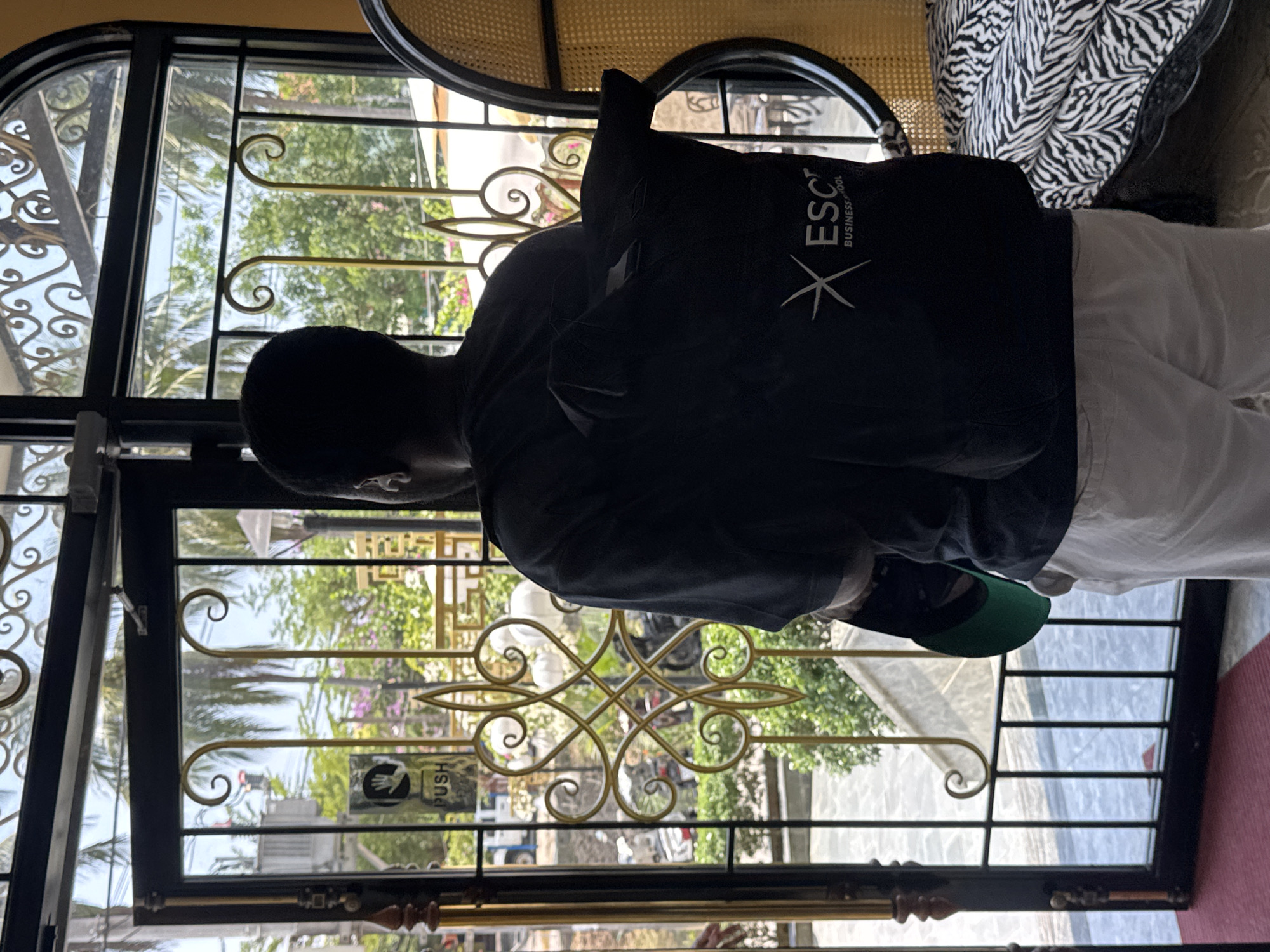 A man wearing a black shirt and a green baseball cap is standing in front of an iron gate. He has a black backpack with white lettering on his back. The gate has a "PUSH" sign and decorative gold metal designs. Outside the gate, vegetation and city infrastructure can be seen. This scene is likely to be in Đà Nẵng, Vietnam.