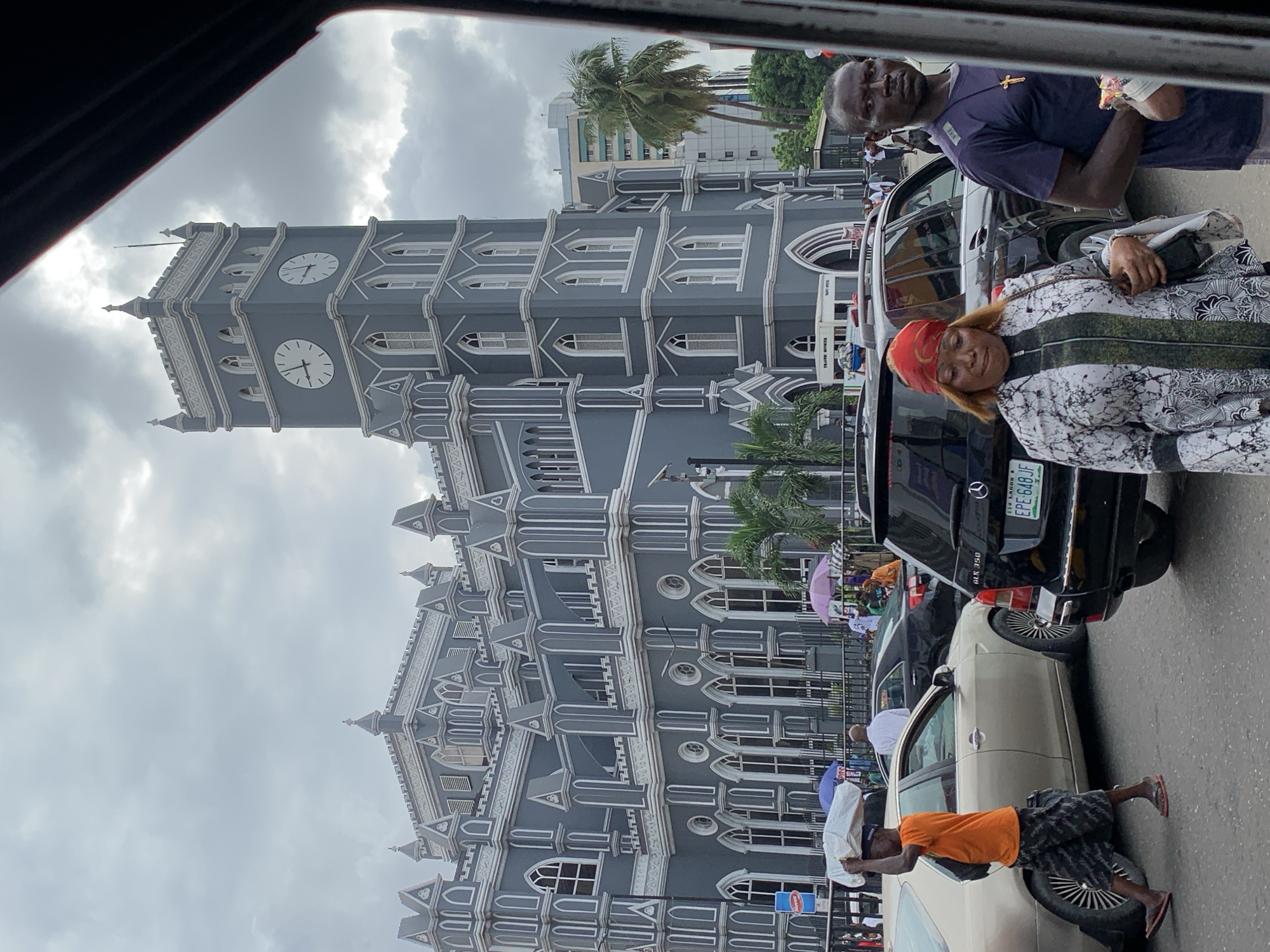An outdoor scene in Lagos, Nigeria, features a bustling street alongside a grand, grey and white Gothic Revival building, likely a cathedral, with a prominent clock tower. The sky is overcast and cloudy.
The building is intricately detailed with pointed arches, buttresses, and small spires, typical of the architectural style. The clock tower has multiple clock faces, with two visible ones showing approximately 11:30 or 11:40. Palm trees rise behind the building and in the background, near other modern structures.
In the foreground, several people and cars are visible. On the right, a dark Mercedes GLK 350 is parked. Standing next to it is a man in a dark blue/purple t-shirt with a gold cross necklace and a white Nike logo on his sleeve, looking towards the viewer. Below him, leaning against the same car, a woman wears a white and black patterned long-sleeved dress and a reddish-orange headscarf, also looking towards the camera while holding a dark object.
Further right, a man in an orange t-shirt and patterned shorts walks past a lighter-colored car, balancing a white bundle on his head. He wears flip-flops. In the background, other pedestrians are seen, some holding colorful umbrellas (purple and blue), suggesting protection from sun or light rain.
Visible text includes the license plate "EPE-648 JF" with "LAGOS" on the Mercedes GLK 350, and the "Nike" logo on the man's shirt. The overall scene depicts a typical street environment in a large city, with people going about their day near a significant landmark.